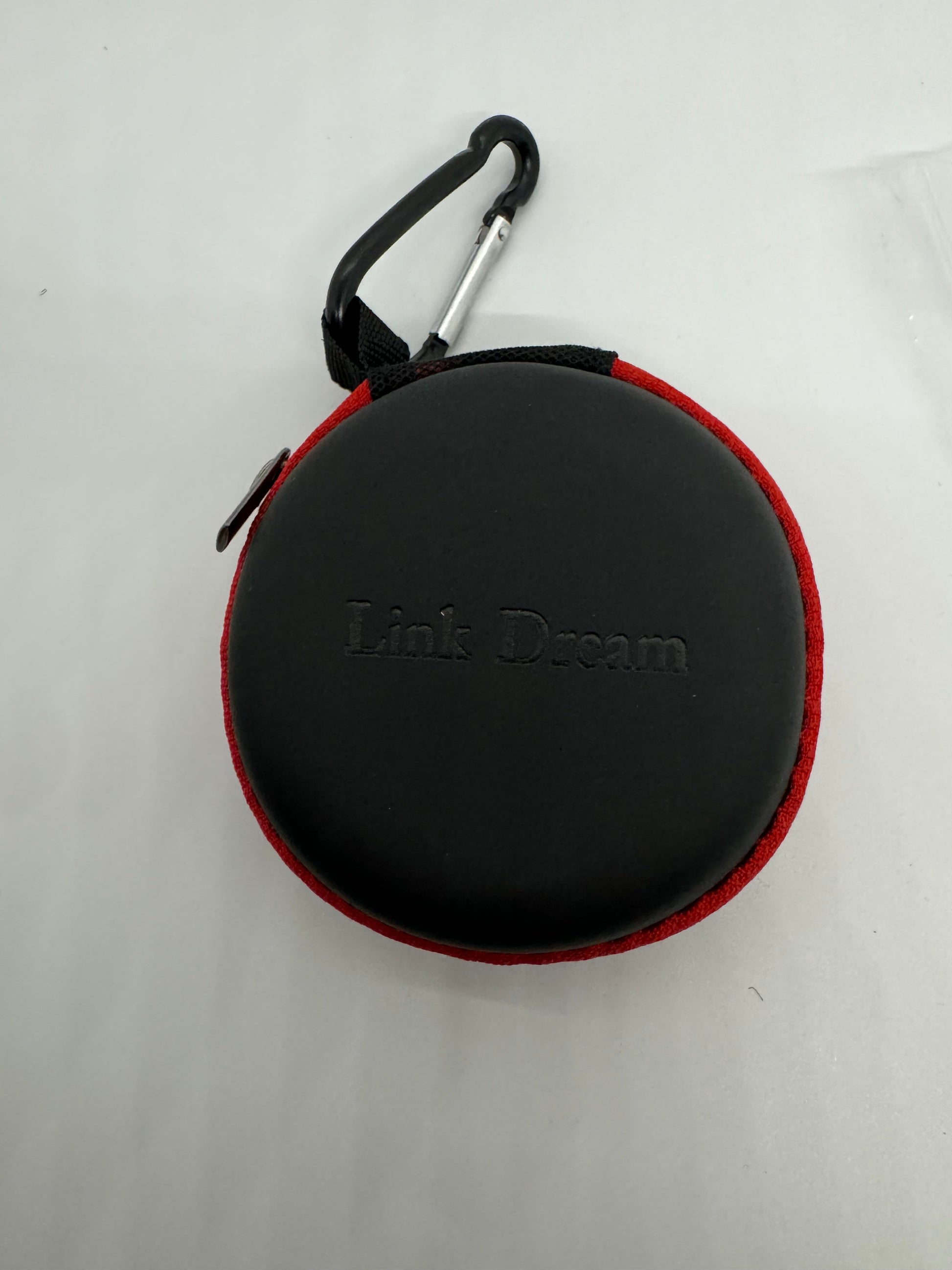 The picture shows a round, black case. The case has a red border and a zipper with a small red pull tab. Attached to the case is a black carabiner clip with a silver gate. The carabiner is attached to the case through a black strap. The background is plain white.