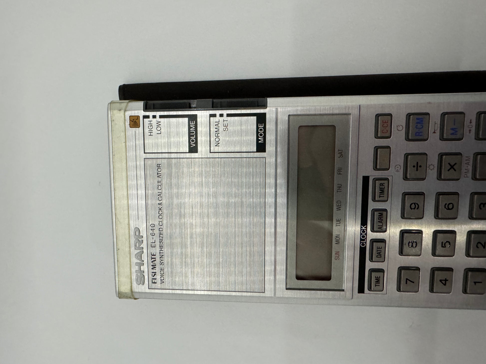The picture shows a Sharp electronic organizer lying on a surface. The organizer has a silver color with a rectangular shape. On the left side, there is a speaker with a volume control slider above it labeled "HIGH" at the top and "LOW" at the bottom. The speaker has horizontal lines across it. On the right side of the organizer, there is a keypad with various buttons. Above the keypad, there is a rectangular screen which seems to be off as nothing is displayed on it. Above the screen, there is