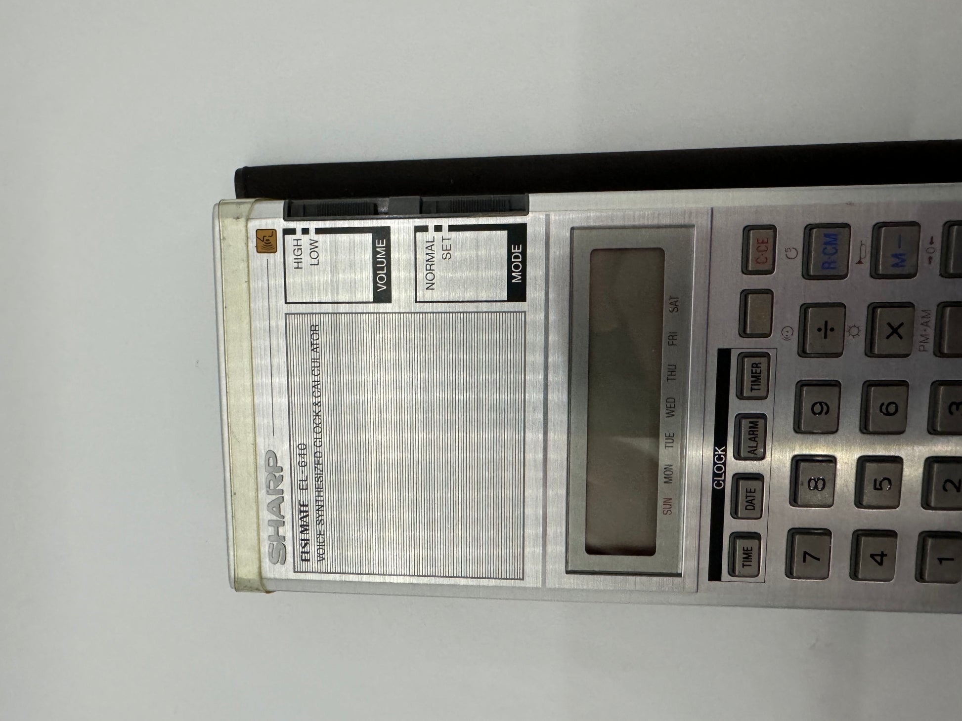 The picture shows a Sharp electronic organizer lying on a surface. The organizer has a silver color with a rectangular shape. On the left side, there is a speaker with a volume control slider above it labeled "HIGH" at the top and "LOW" at the bottom. The speaker has horizontal lines across it. On the right side of the organizer, there is a keypad with various buttons. Above the keypad, there is a rectangular screen which seems to be off as nothing is displayed on it. Above the screen, there is