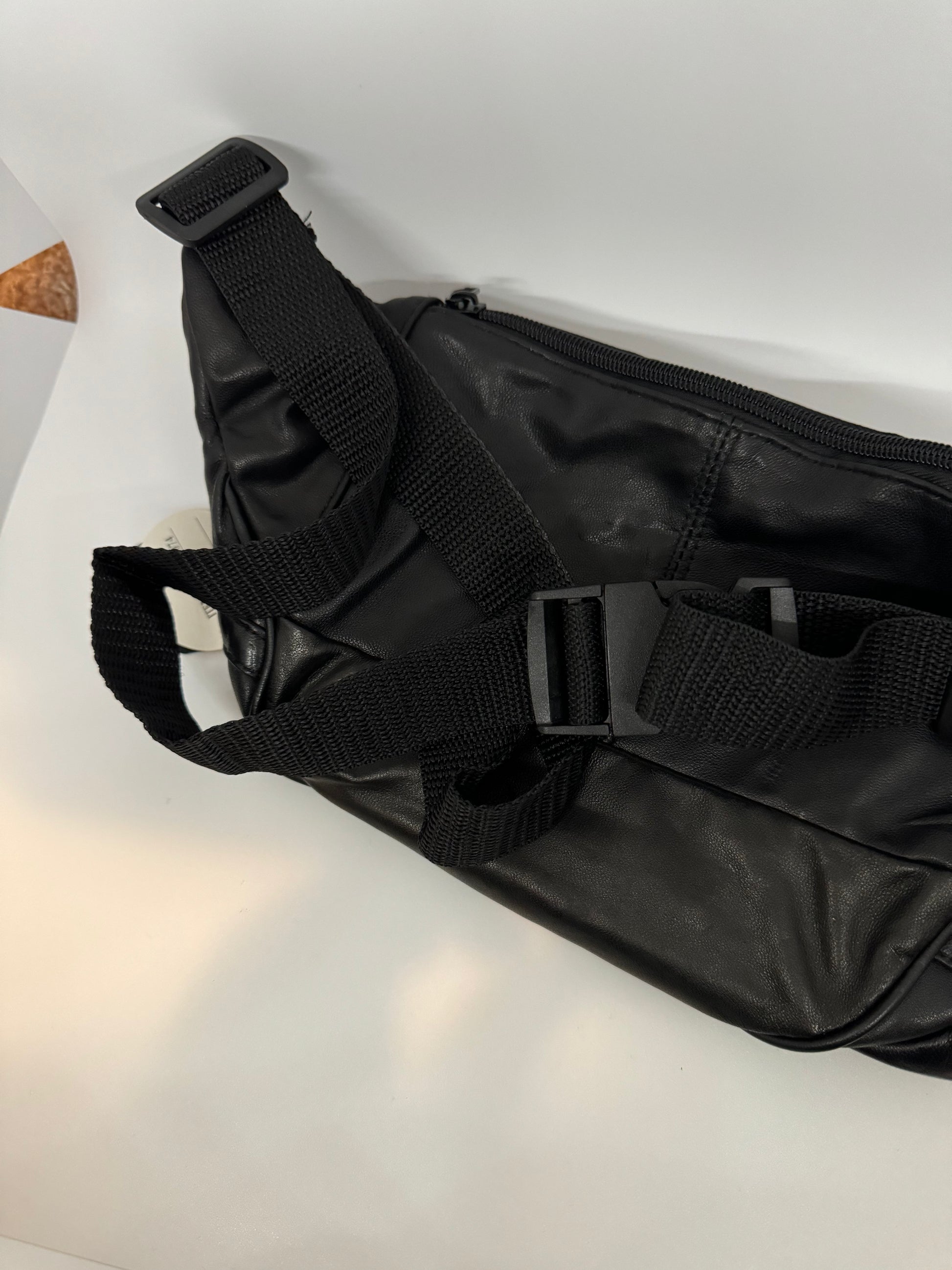 The picture shows a black bag, possibly made of leather or a similar material. The bag has a zipper on top. There is a black woven strap attached to the bag, which looks like it's adjustable with a plastic buckle. The background is white.