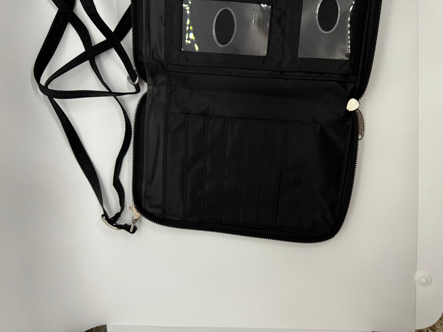 The picture shows a black wallet or organizer with a strap. The wallet is open and has multiple compartments. On the left side, there is a strap that is likely used to carry or secure the wallet. The strap is black and has a clip at the end. On the right side of the wallet, there are several vertical slots that can be used to store cards or other flat items. Above these slots, there is a zippered compartment. The zipper has a silver pull tab. The wallet also has a clear plastic section with three