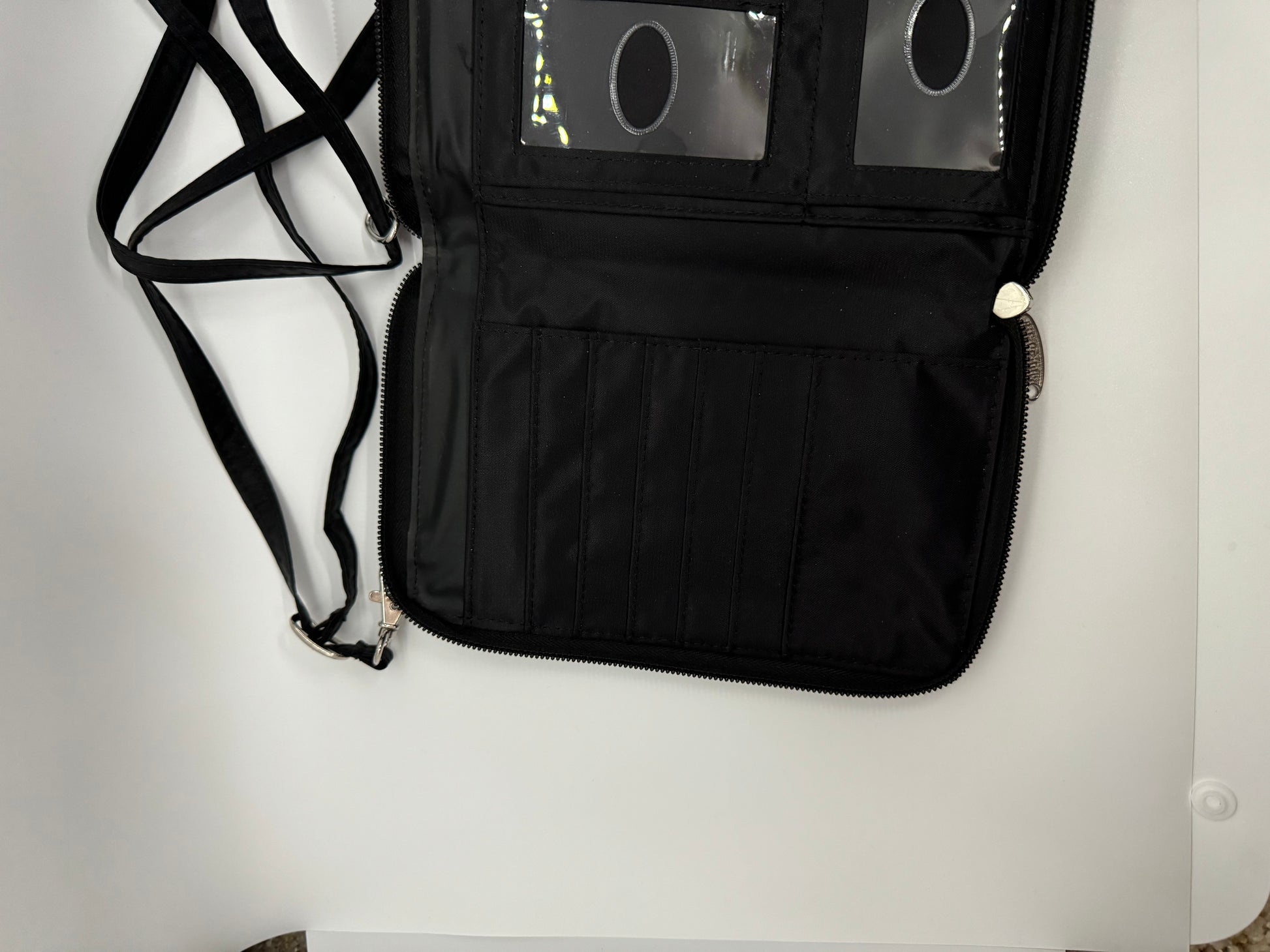 The picture shows a black wallet or organizer with a strap. The wallet is open and has multiple compartments. On the left side, there is a strap that is likely used to carry or secure the wallet. The strap is black and has a clip at the end. On the right side of the wallet, there are several vertical slots that can be used to store cards or other flat items. Above these slots, there is a zippered compartment. The zipper has a silver pull tab. The wallet also has a clear plastic section with three