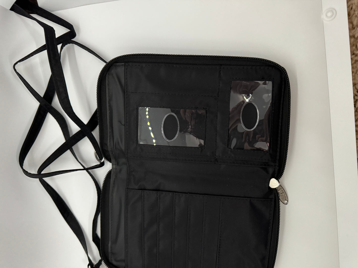 The picture shows a black wallet or organizer with a strap. The wallet is open and lying flat on a white surface. The left side of the wallet has a zipper running along the edge. The right side has multiple compartments, including a clear plastic window, possibly for an ID card, and several slots for cards or other small items. The strap is black and appears to be made of a fabric material. It is looped and lying next to the wallet. The zipper pull is silver and has a decorative shape.