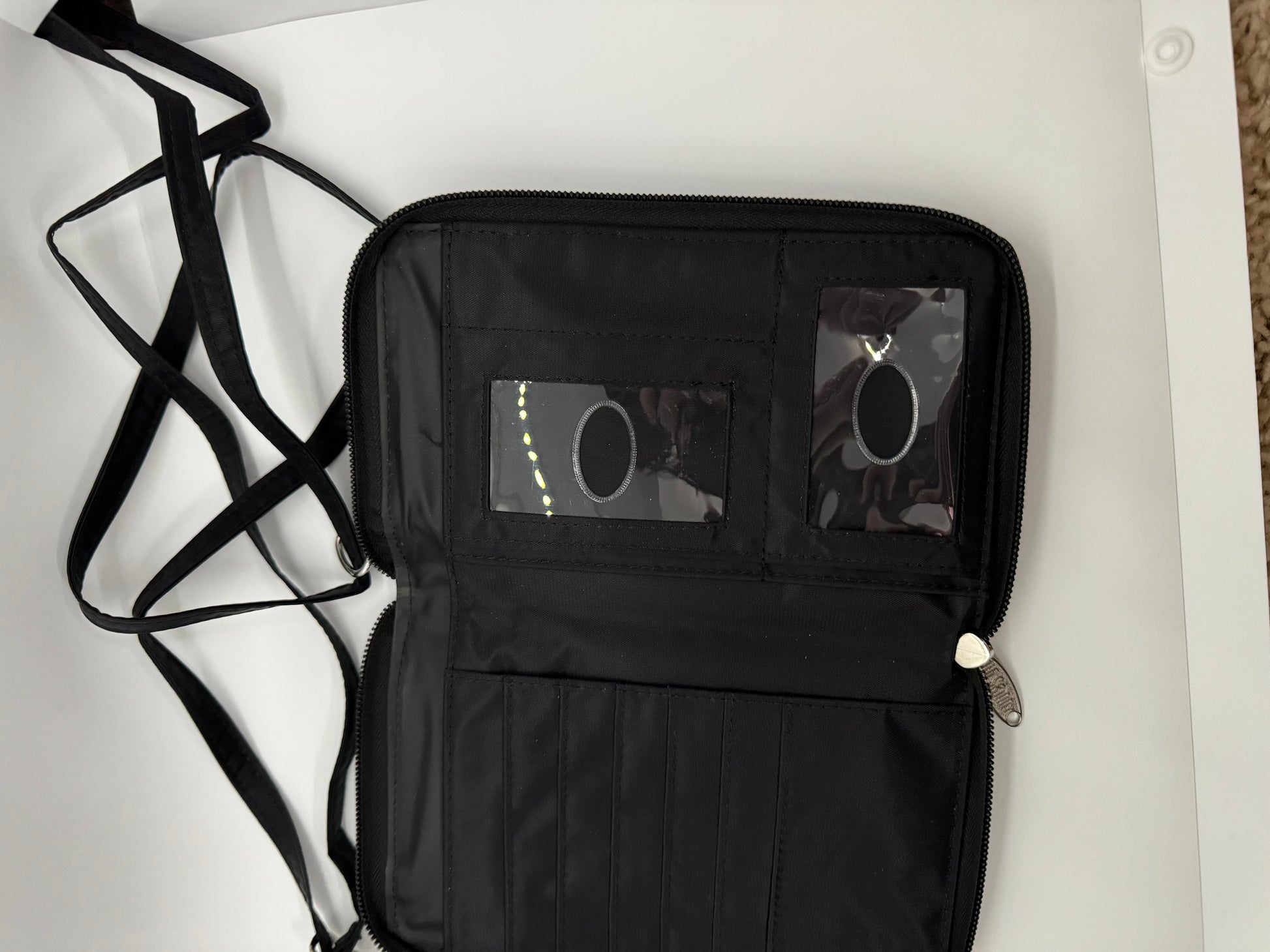 The picture shows a black wallet or organizer with a strap. The wallet is open and lying flat on a white surface. The left side of the wallet has a zipper running along the edge. The right side has multiple compartments, including a clear plastic window, possibly for an ID card, and several slots for cards or other small items. The strap is black and appears to be made of a fabric material. It is looped and lying next to the wallet. The zipper pull is silver and has a decorative shape.