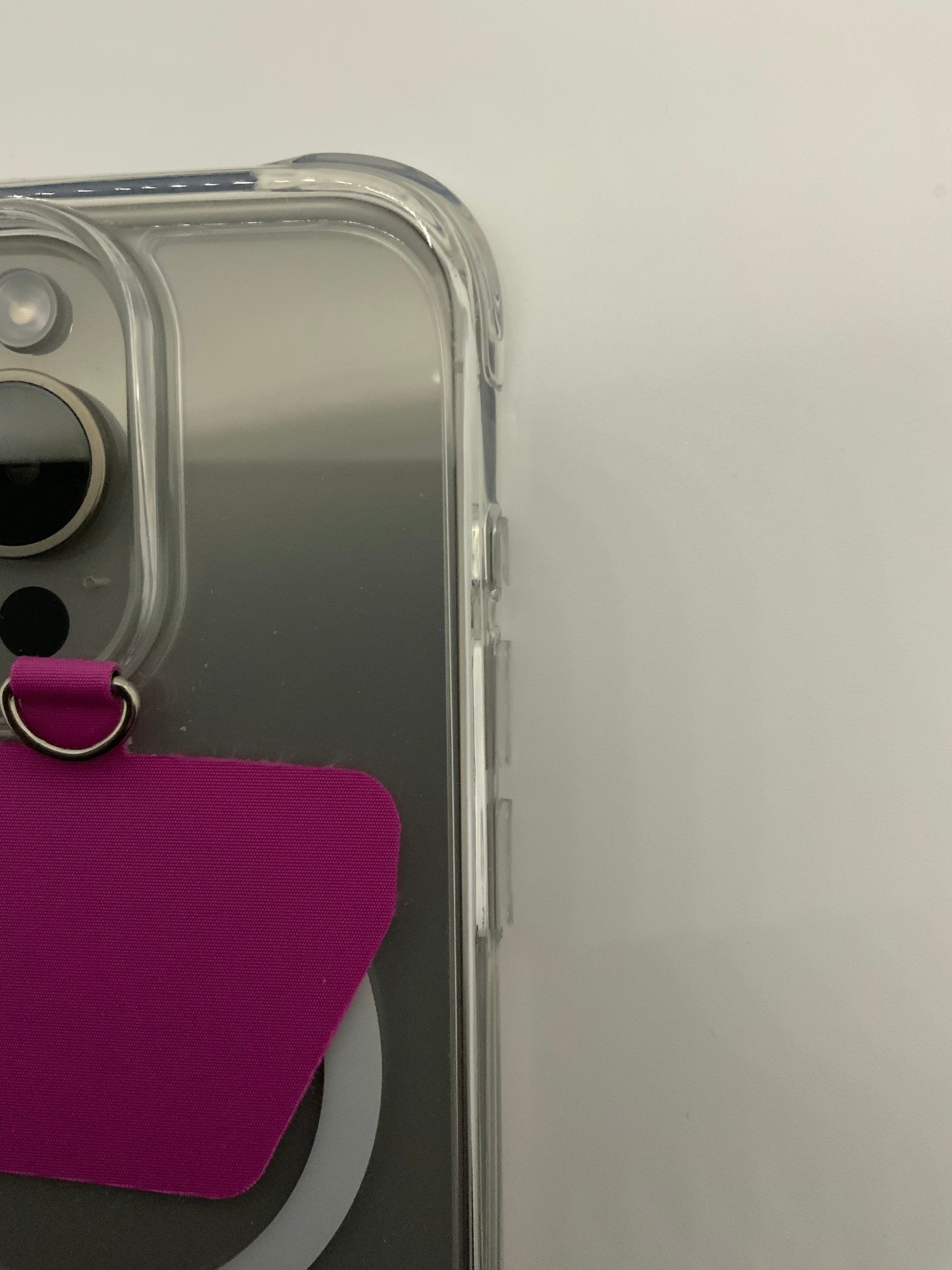 Be My AI: The picture shows a close-up of the corner of a clear phone case that is on a phone. The phone case has a transparent, plastic material and you can see the side of the phone through it. The phone has a camera in the top left corner. There is also a purple tag with a metal ring attached to the back of the phone, which is visible through the clear case. The background is plain and white.