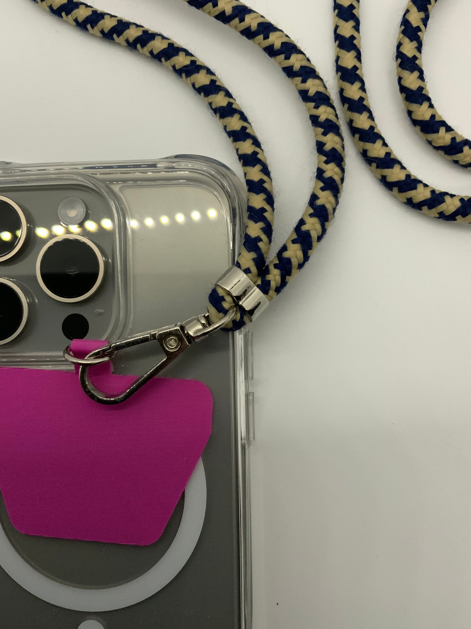 Be My AI: The picture shows a clear phone case with a lanyard attached to it. The phone case appears to be for an iPhone with three camera lenses and a flash. The lanyard is made of a braided fabric in a pattern of alternating blue and beige colors. The lanyard is attached to the phone case through a silver metal clasp. There is also a pink card or tag attached to the back of the phone case. The background of the picture is white.