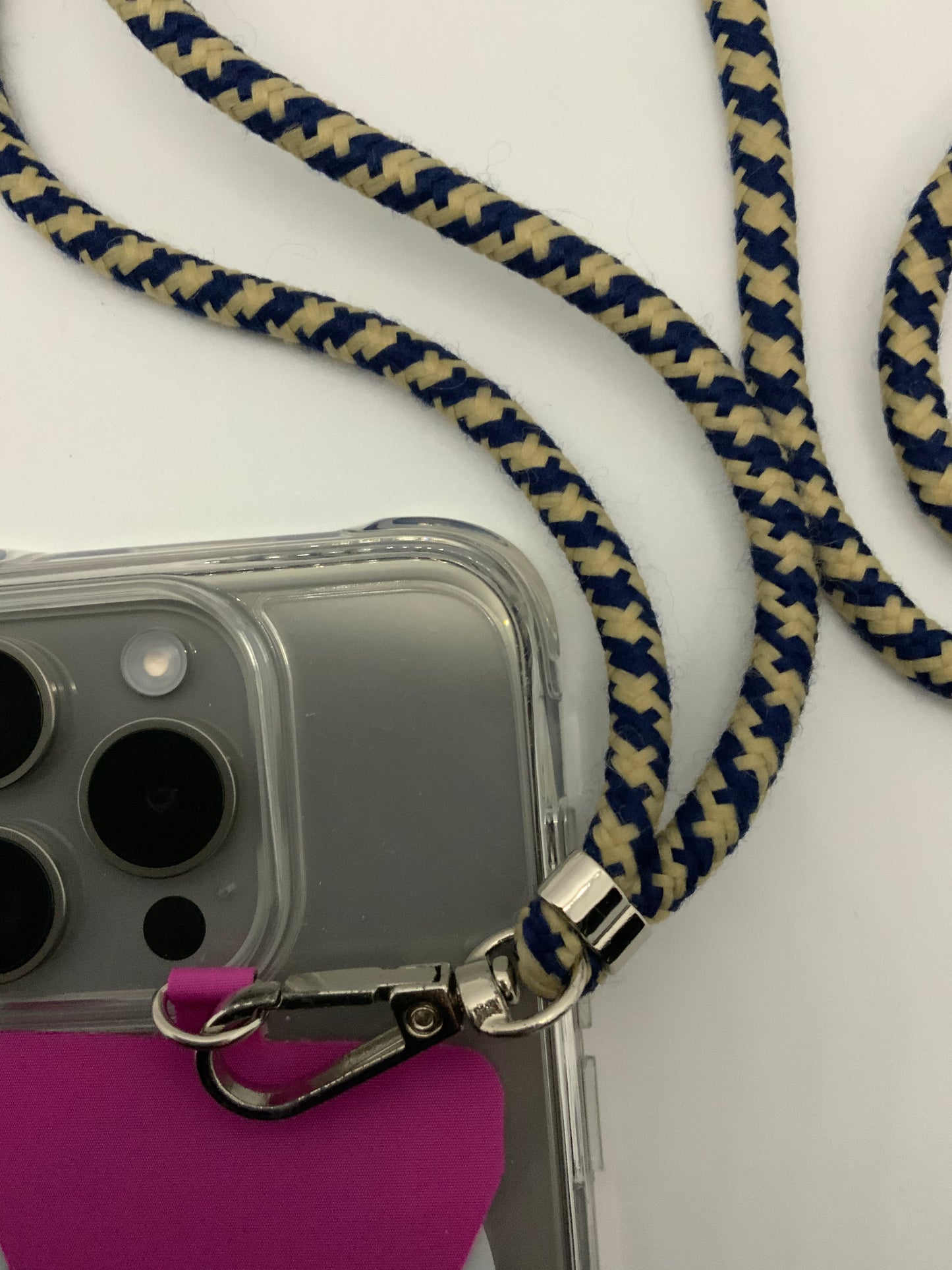 Be My AI: The picture shows a close-up of a woven lanyard and a clear phone case. The lanyard is made of a fabric material and has a pattern of alternating navy blue and beige colors. The lanyard is attached to a silver metal clasp, which is hooked onto the corner of a clear phone case. The phone case has a pink card holder on the back and is designed for a phone with three camera lenses and a flash. The background of the picture is white.