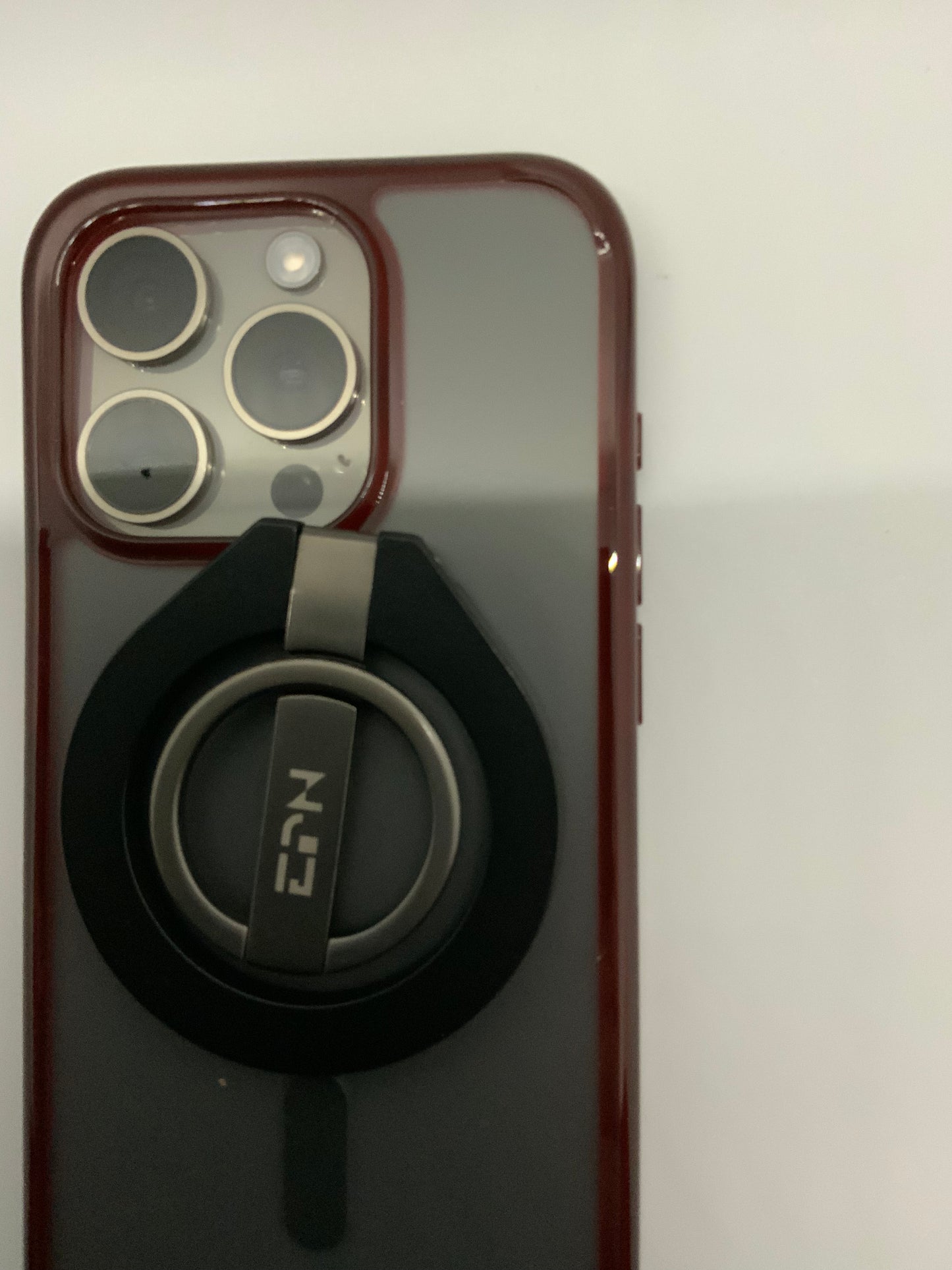 Be My AI: The picture shows a close-up of the back of a smartphone with a red case. The camera module is visible in the top left corner of the phone. It has three circular lenses and a flash arranged in a square shape. Below the camera module, there is a black ring holder attached to the phone case. The ring holder has a silver-colored metal ring and a base with the letters "NCL" written on it. The background is plain white.