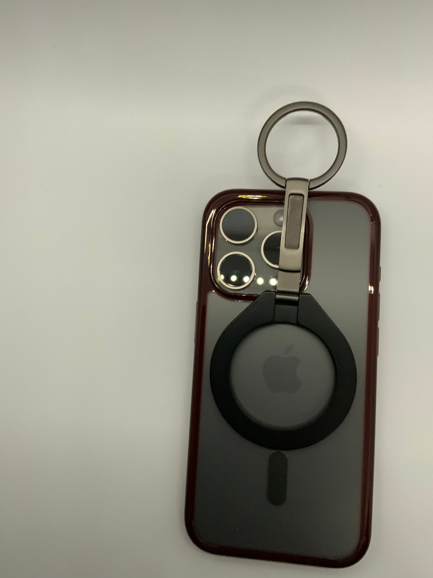 Be My AI: The picture shows a smartphone with a case and an attached accessory. The smartphone case is a deep red color and has a cutout on the back to show the Apple logo, which is centered. Above the Apple logo, there is a large cutout for the camera and flash which are in a square configuration with rounded corners. The camera cutout has three circles for the lenses and a smaller circle for the flash.

Attached to the top of the smartphone case is a circular accessory with a ring. The accessory is attach