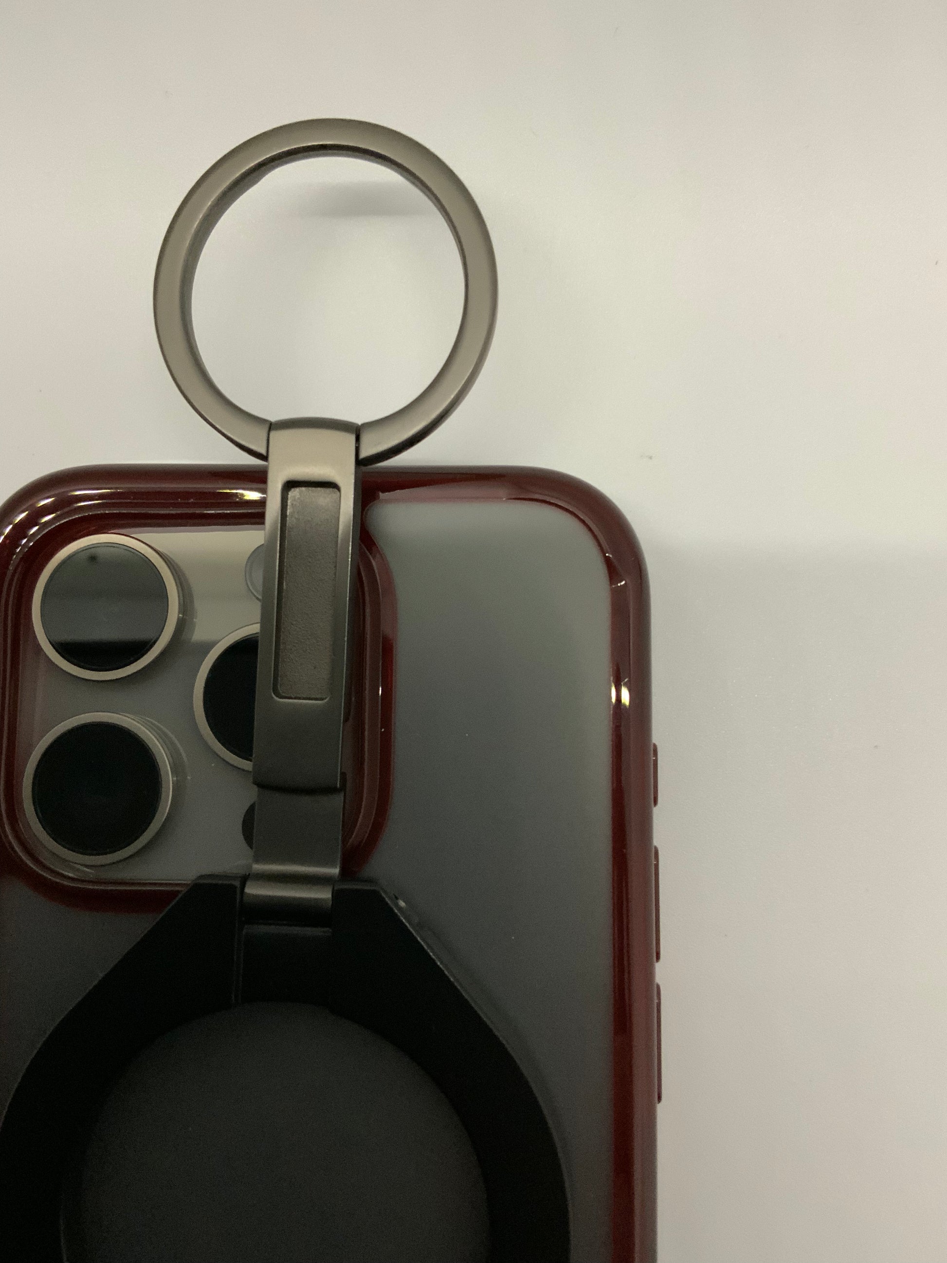 Be My AI: The picture shows a close-up of what appears to be a phone case with an attached ring holder. The phone case is a deep red color with a cutout for the camera and flash on the back of the phone. The ring holder is attached to the back of the case and is made of metal. It has a circular ring at the top, which is attached to a base that is adhered to the phone case. The ring is in an upright position and can likely be used to hold the phone securely with a finger through the ring or as a stand to pro
