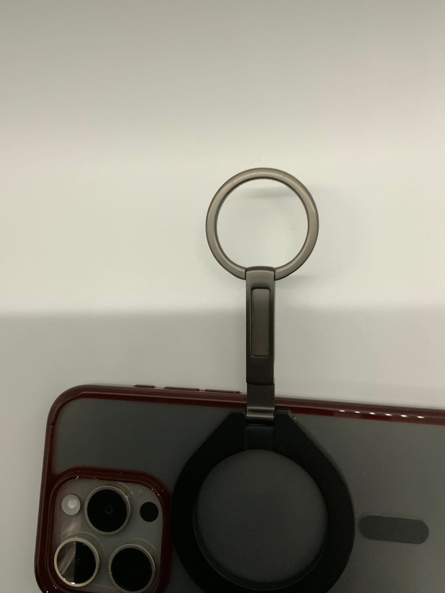 Be My AI: The picture shows a part of a smartphone with a red case and a ring holder attached to it. The smartphone case has a cut-out for the camera, which has three lenses and a flash. The ring holder is attached to the back of the smartphone case. It has a metal ring and a base. The ring is in the upright position and can be used to hold the phone securely or as a stand. The background is plain and light-colored.