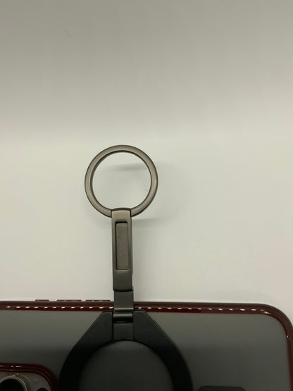 Be My AI: The picture shows a ring holder attached to the back of a device, possibly a smartphone. The ring holder is in the center of the image. It has a metallic ring attached to a base. The base is rectangular and seems to be attached to the device. The device has a maroon border with stitching details. The background is plain and light-colored.