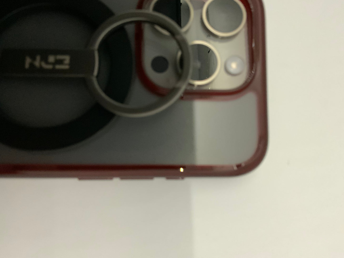 Be My AI: The picture shows a close-up of a few objects. On the left side, there is a black object with a circular shape and a metal ring attached to it. The metal ring has a small rectangular tag with the letters "N" and "Z" on it. On the right side, there is a part of a red phone case with a camera cutout. The camera cutout has three circular holes and one oval hole. The background is white.