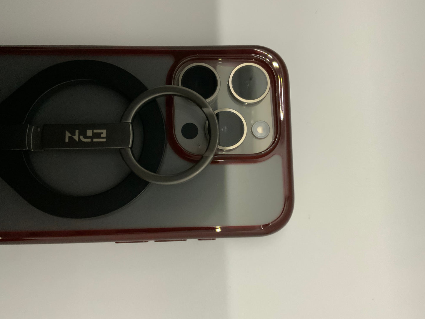 Be My AI: The picture shows a close-up of a few objects on a white surface. On the right side, there is a part of a phone with a red case. The phone has a camera module with three lenses and a flash. On the left side, there is a black object with a circular base and a metal ring attached to it. The metal ring is attached to a black rectangular piece with the letters "N" and "E" visible on it, suggesting that it might be a brand or model name. The objects are placed close to each other.