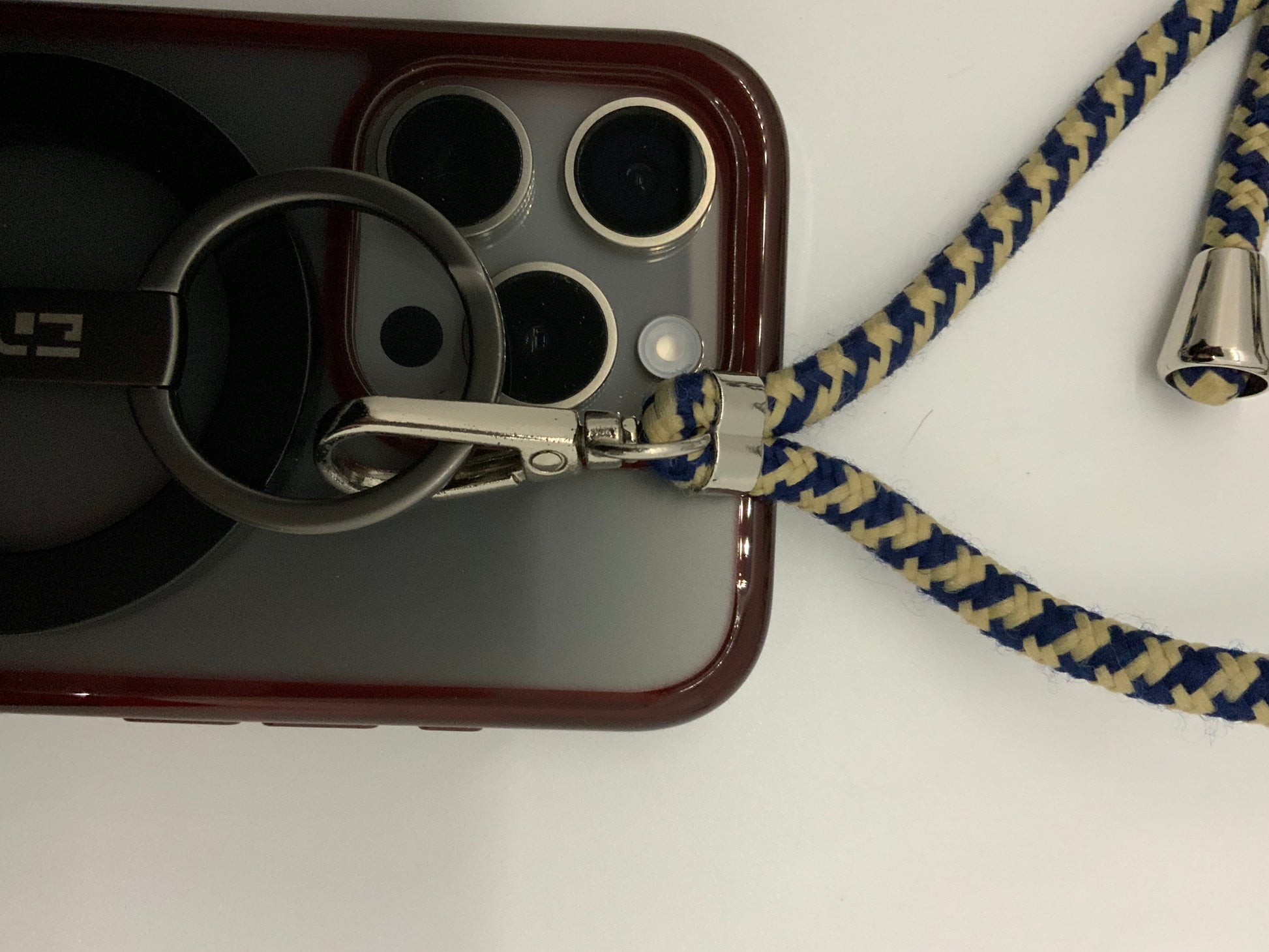 Be My AI: The picture shows a close-up of a few objects. There is a phone with a red case that has a camera with three lenses and a flash in a square arrangement. Attached to the phone case is a black carabiner with a silver clip. The carabiner has the letters "LE" on it. There is also a blue and beige braided cord attached to the silver clip. The cord has a silver bell at the end. The background is white.