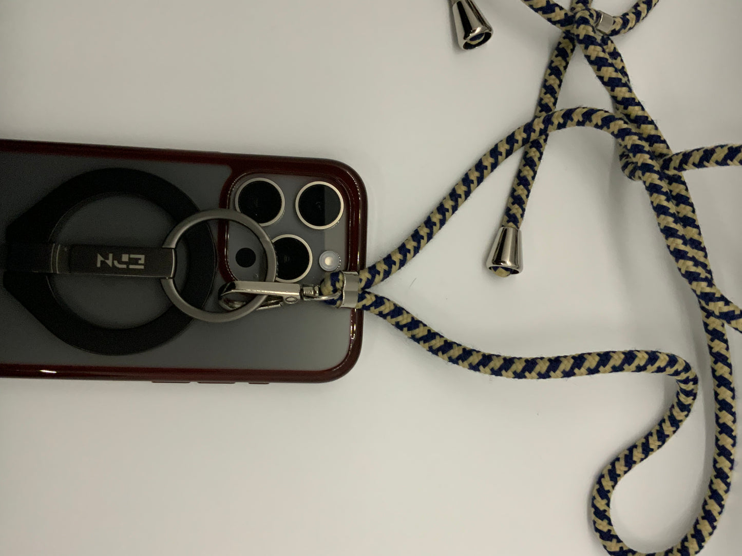 Be My AI: The picture shows a phone case with a strap attached to it. The phone case is burgundy in color and has a cutout for the camera and flash. Attached to the case is a circular ring holder with the letters "N2J" on it. The ring holder is attached to a strap that is made of a fabric material. The strap is braided and has a pattern of blue and beige colors. The strap also has metal aglets at the ends. The background of the picture is white.