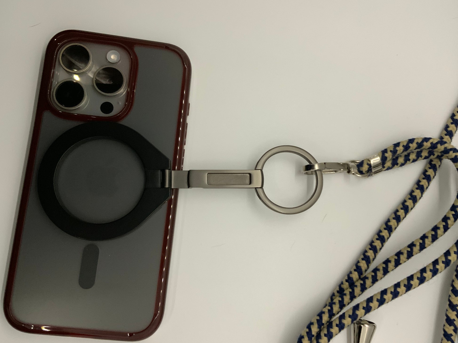 Be My AI: The picture shows a smartphone with a case on it. The case is dark red with a black circular attachment on the back, possibly for grip or as a stand. The phone's camera is visible at the top left corner of the case, with three lenses and a flash.

There is also a lanyard attached to the case. The lanyard has a metal clip and a keyring attached to it. The strap of the lanyard is made of fabric with a zigzag pattern in dark blue and beige colors.

The background is plain white.