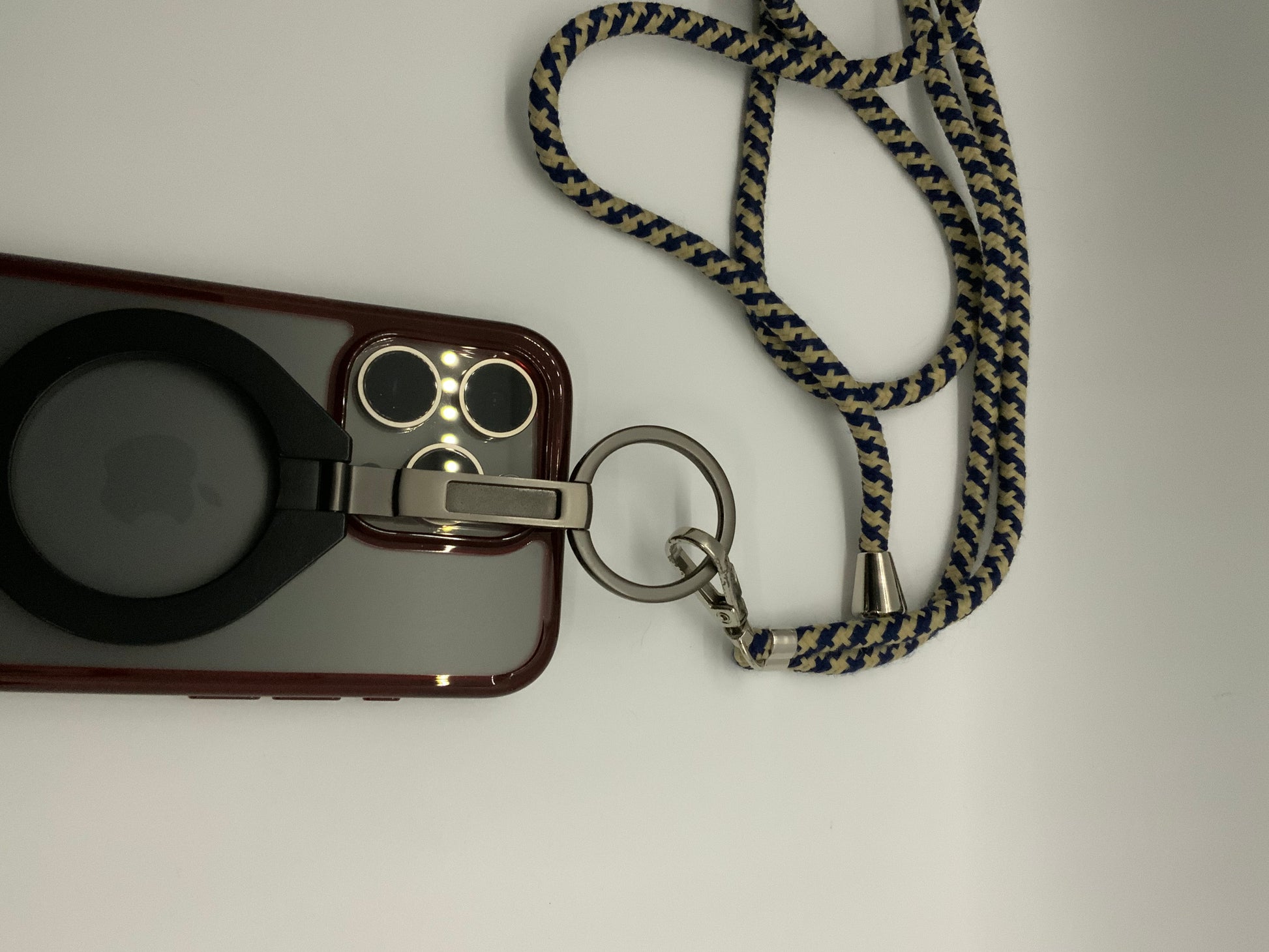 Be My AI: The picture shows a phone with a case and a lanyard attached to it. The phone case is transparent with a black circular ring in the middle, possibly for holding or standing the phone. The case has a maroon border. The camera cut-out on the case is square with rounded edges and shows three camera lenses and a flash. 

Attached to the case is a lanyard with a metal clip. The lanyard is made of fabric and has a zigzag pattern in dark blue and beige colors. The metal clip is silver and is attached to 