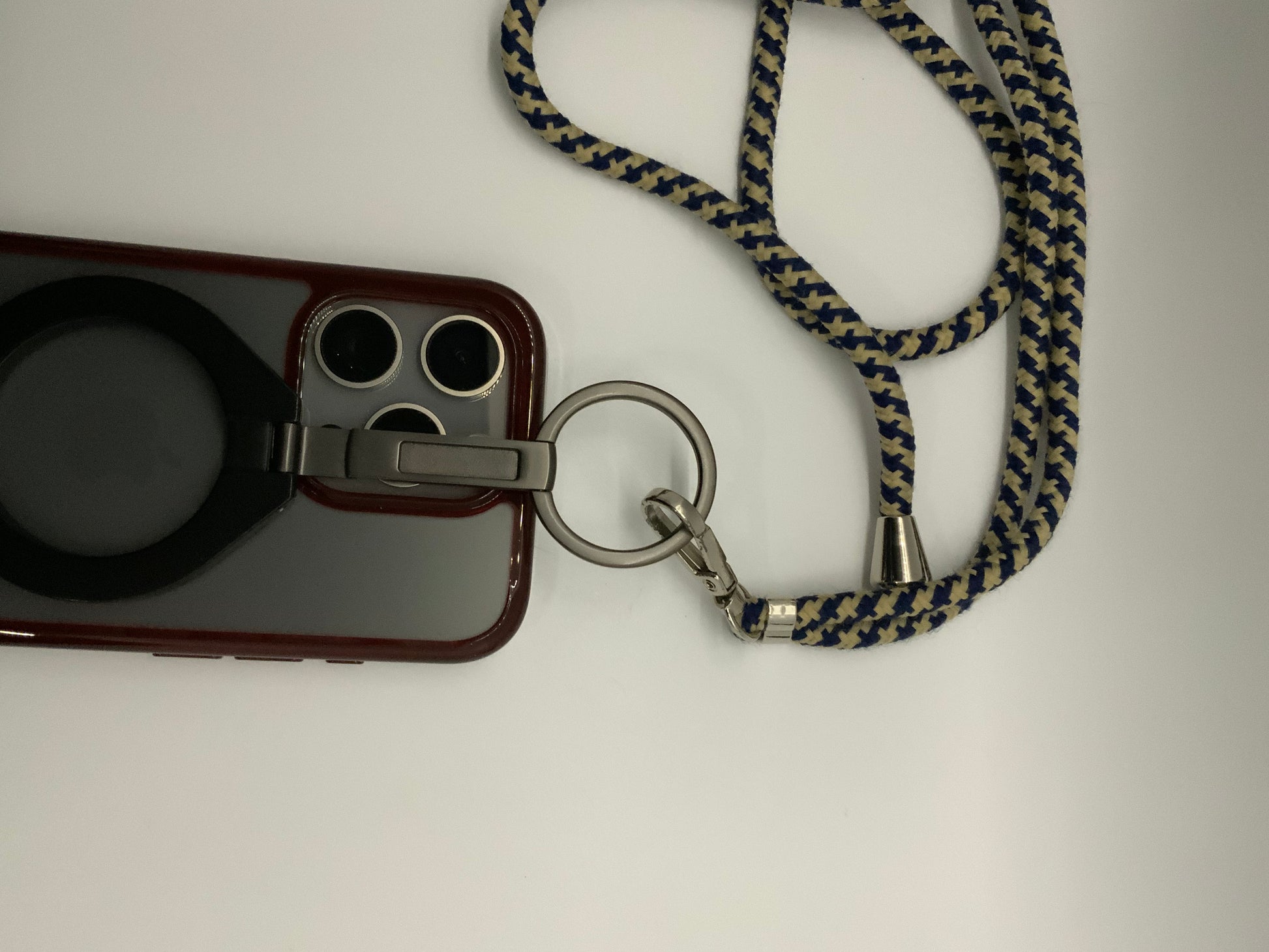 Be My AI: The picture shows a phone case with a strap. The phone case is a deep red color and has a circular cutout on the back, possibly for a logo. There are also three circular cutouts at the top left corner of the case, likely for the camera lenses. Attached to the case is a metal ring with a strap. The strap is made of fabric and has a zigzag pattern in dark blue and beige colors. The strap is looped and appears to be long enough to wear around the neck. There is also a small metal bell attached to the