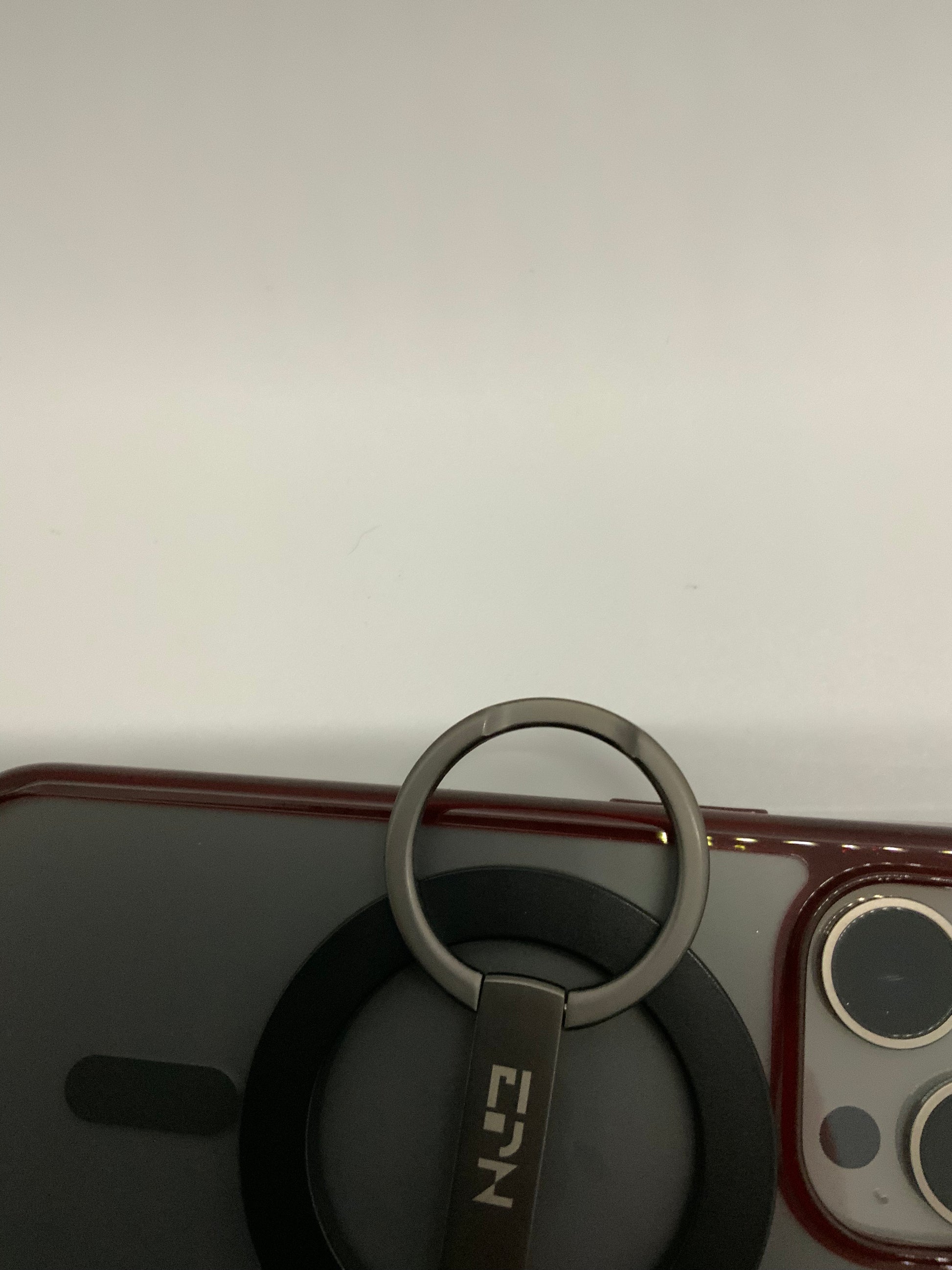 Be My AI: The picture shows a close-up of what appears to be the back of a smartphone with a ring holder attached to it. The smartphone has a red border and a dark-colored back. On the top right corner of the smartphone, there are two camera lenses and a flash within a rectangular section. The ring holder is attached to the back of the smartphone and has a dark circular base with a metal ring. The metal ring is attached to a small rectangular piece that has the letters "EIN" written on it. The background is