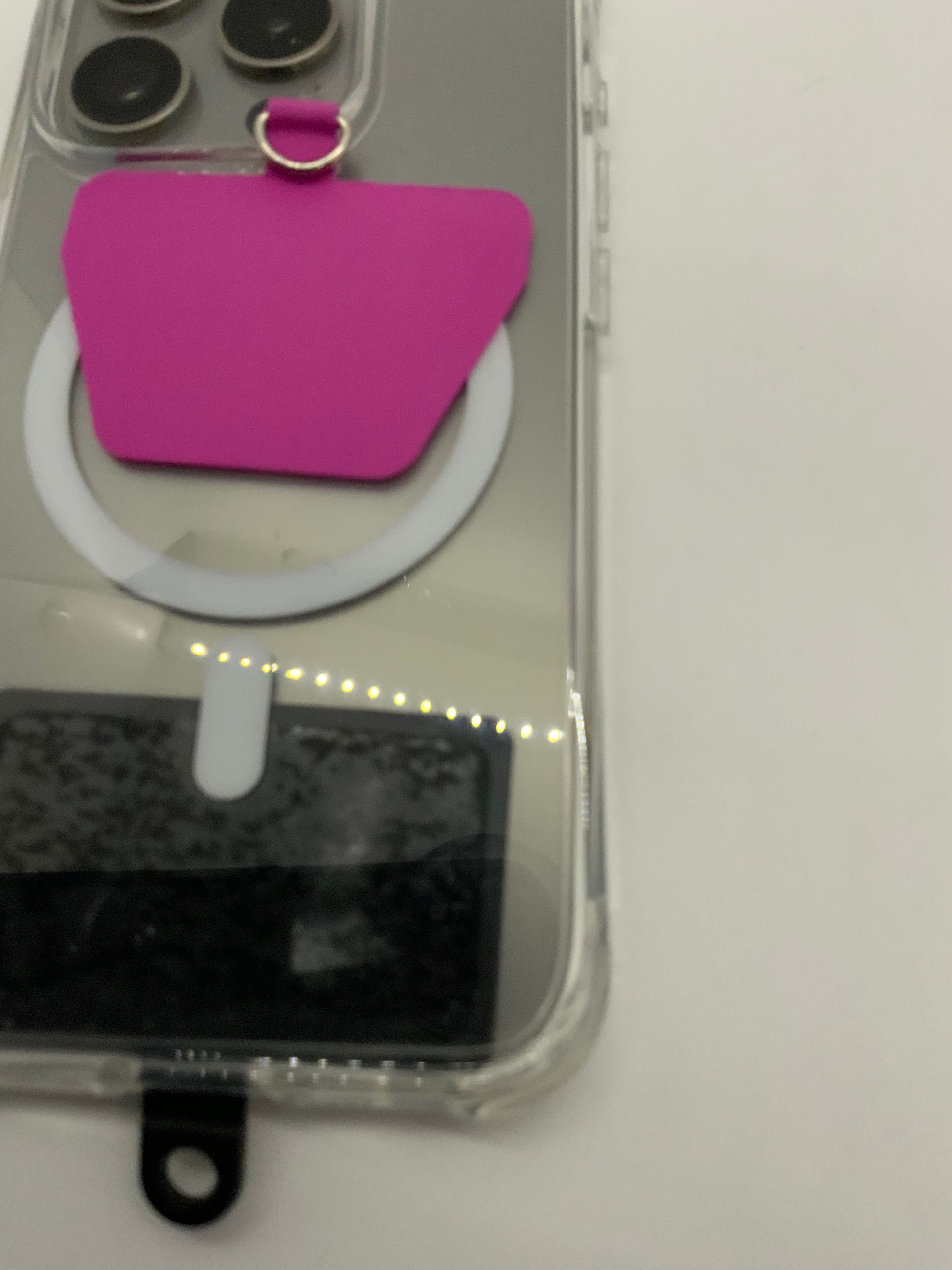 The picture shows a close-up of a few objects on a white surface. There is a bright pink, flat object with a small golden ring attached to the top. Below it, there is a white circular ring. To the left, there are four black circular objects in a transparent packaging. Below the white ring, there is a rectangular object with a black and grey pattern, which is also in transparent packaging. The image is slightly blurred.