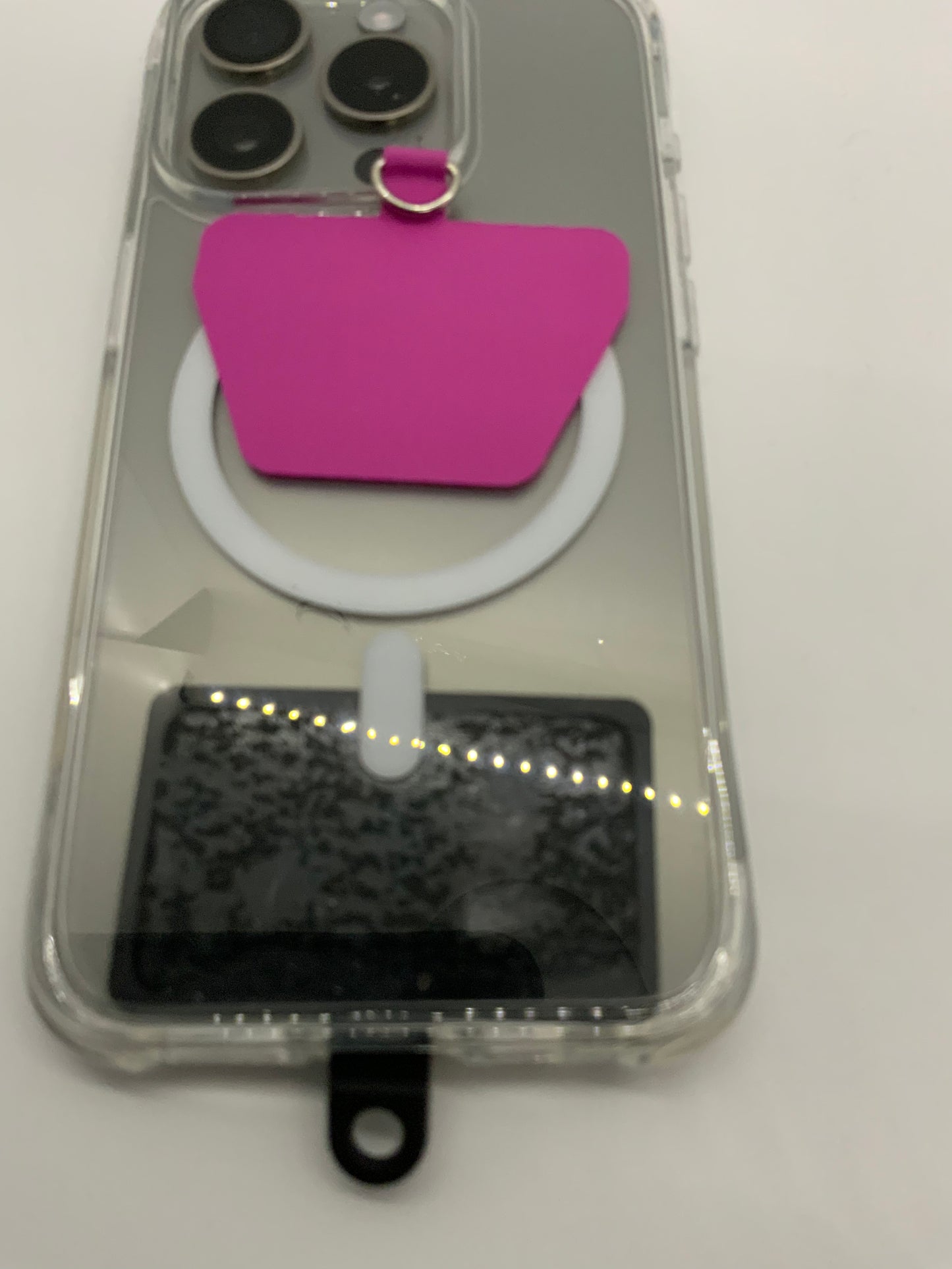 The picture shows a close-up of the back of a smartphone with a clear case. The phone has three camera lenses in a triangular arrangement at the top left corner. There is a pink adhesive wallet attached to the back of the case, which is shaped like a small pocket with a ring at the top. Below the wallet, there is a white circular object that seems to be attached to the case. The bottom part of the phone is visible, showing a black strip with a small tab, possibly for opening the case. The backgrou