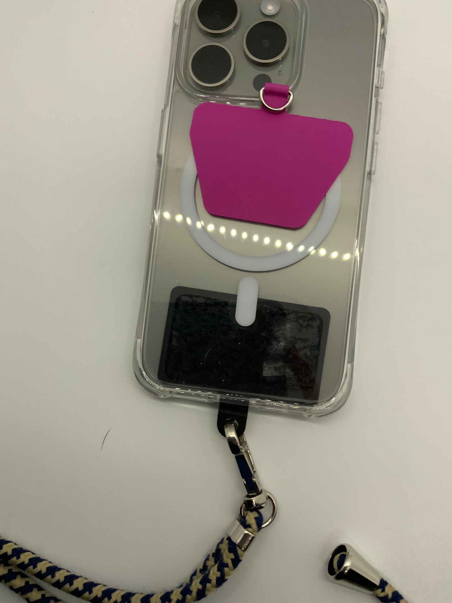 The picture shows a phone with a clear case on a white background. The phone has three camera lenses and a flash in a square arrangement at the top left corner. There is a pink sticky note attached to the back of the phone case, and it has a small metal ring attached to it. Below the sticky note, there is a white circular object with LED lights. The phone case also has a slot for an ID or credit card at the bottom. Attached to the bottom of the phone case is a blue and white braided lanyard with m