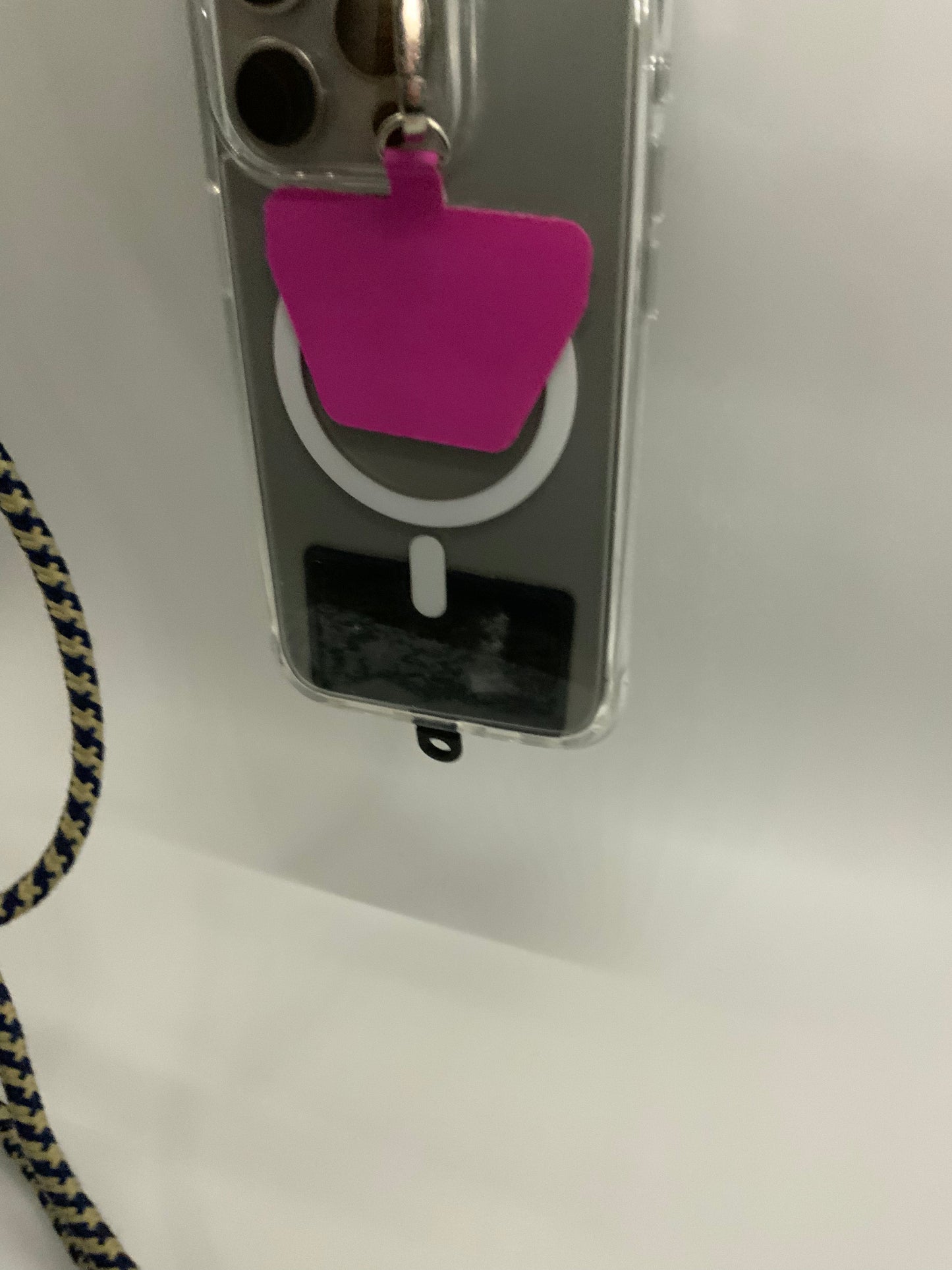 The picture shows a close-up of an object that appears to be a badge holder or a small device. It has a clear plastic casing with a slot at the top where a pink tag is attached through a metal loop. Below the slot, there is a white circular button with a black rectangular area beneath it. The background is plain white and there is a partial view of a cord with blue and yellow patterns at the bottom left corner of the picture.