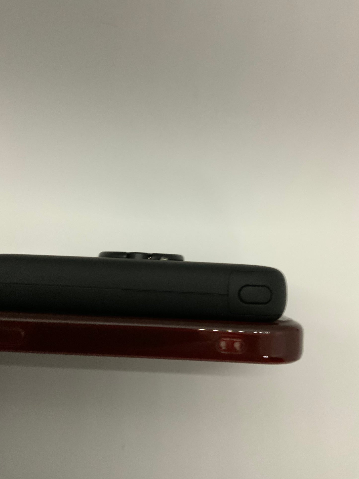 The picture shows two objects that appear to be smartphones or similar devices, stacked on top of each other against a plain light background. The top device is black and has a button on the side. The bottom device is a dark red or maroon color and appears to be slightly thicker than the black one. The bottom device also has a button on the side, and there is a small symbol or logo next to the button. The devices are shown from the side, and the focus is on the edges of the devices.