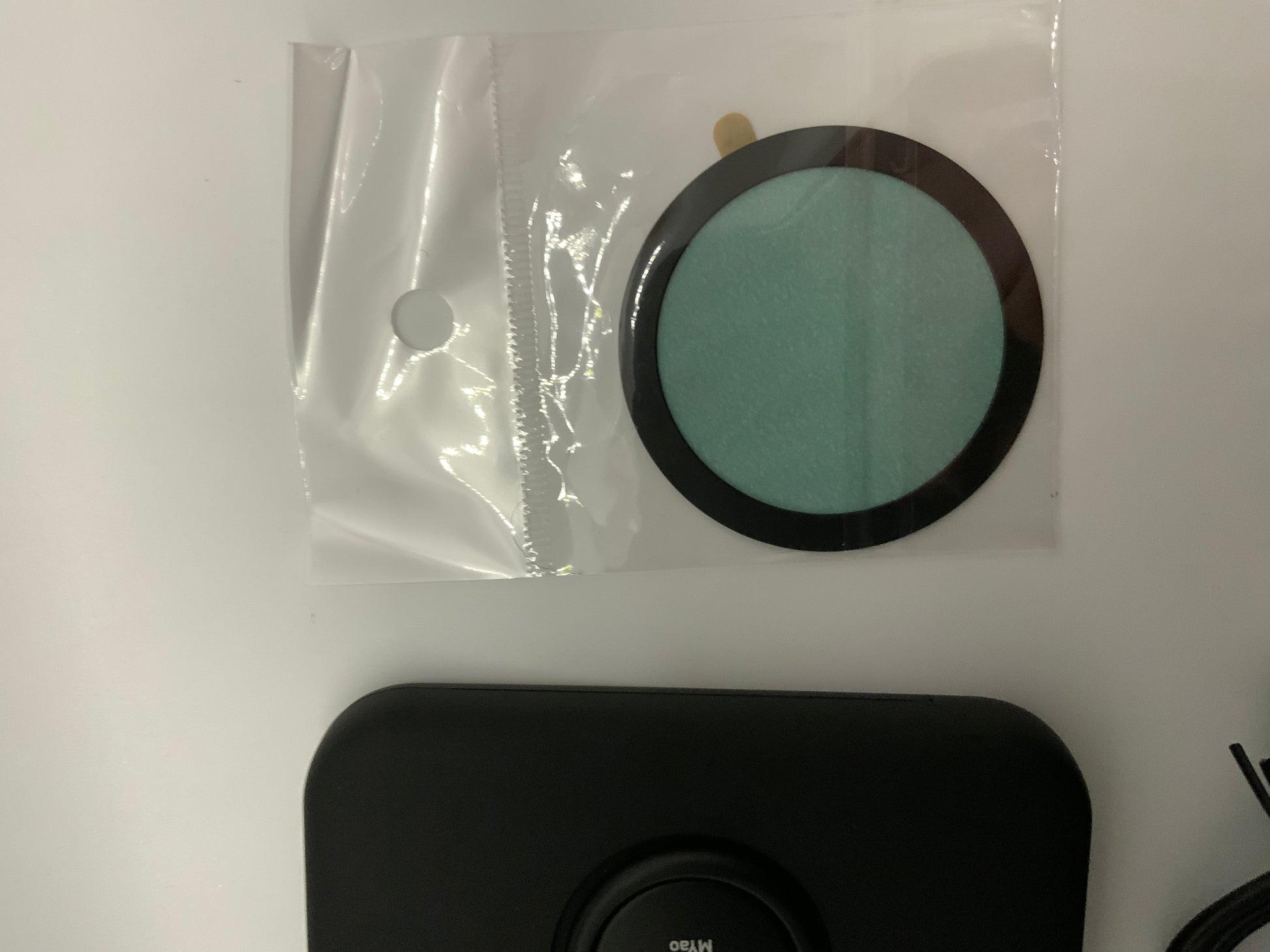 The picture shows three items on a white surface. 1. On the top left, there is a clear plastic bag with a white circular sticker sealing it. The bag is empty and has a glossy finish.2. To the right of the plastic bag, there is a circular object with a black outer ring and a light green center. It appears to be a filter or a pad of some sort. It is also inside a clear plastic bag.3. At the bottom of the picture, there is a black rectangular object with rounded corners. It has a circular inden