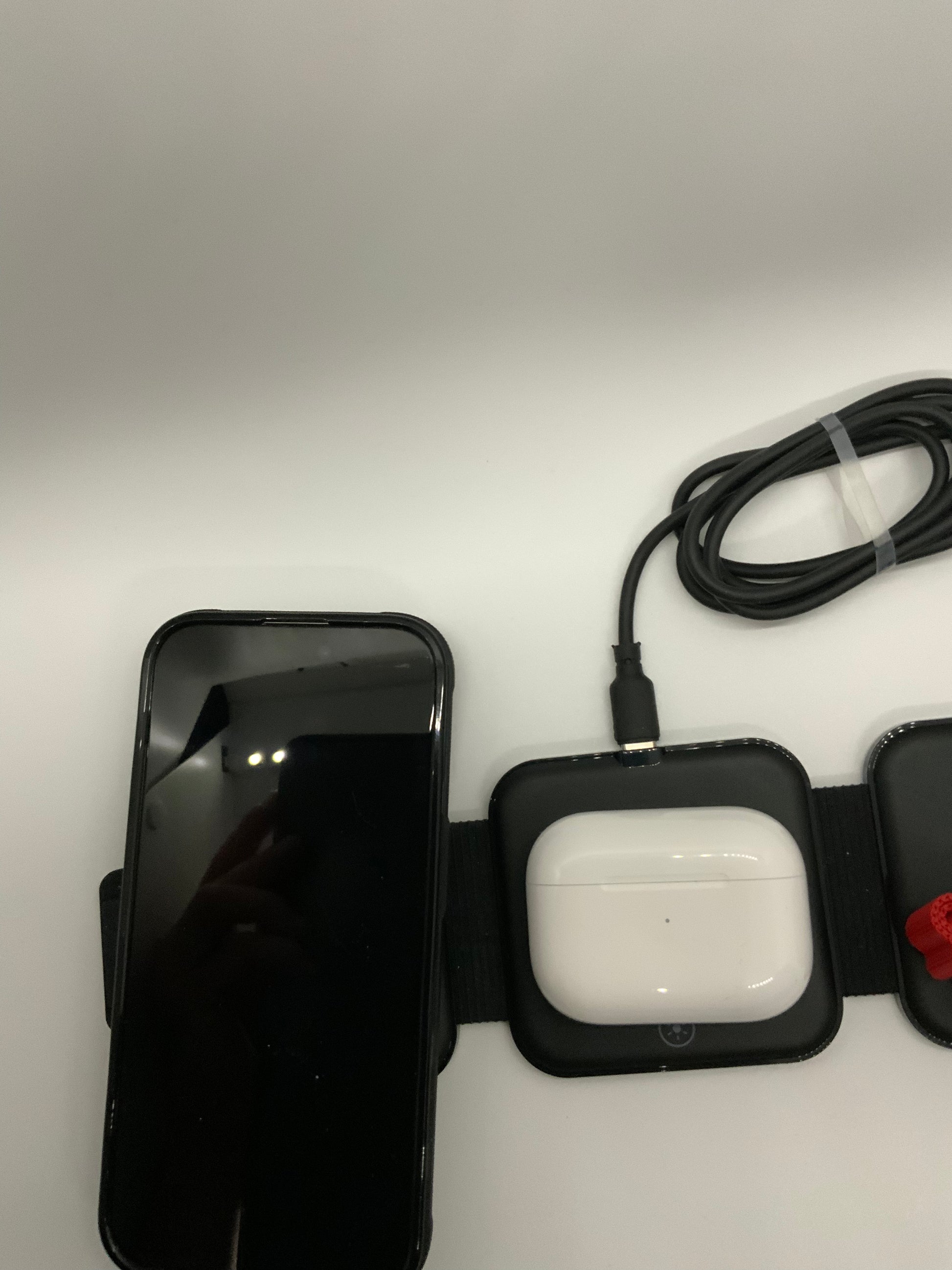 Be My AI: The picture shows a few items placed on a white surface. On the left side, there is a smartphone with a black screen, which seems to be turned off. The phone is in a black case with a strap attached to it. 

On the right side, there is a white case of wireless earbuds placed on a black square pad with a strap. Above the earbuds case, there is a black cable coiled up. The cable has a USB connector on one end.