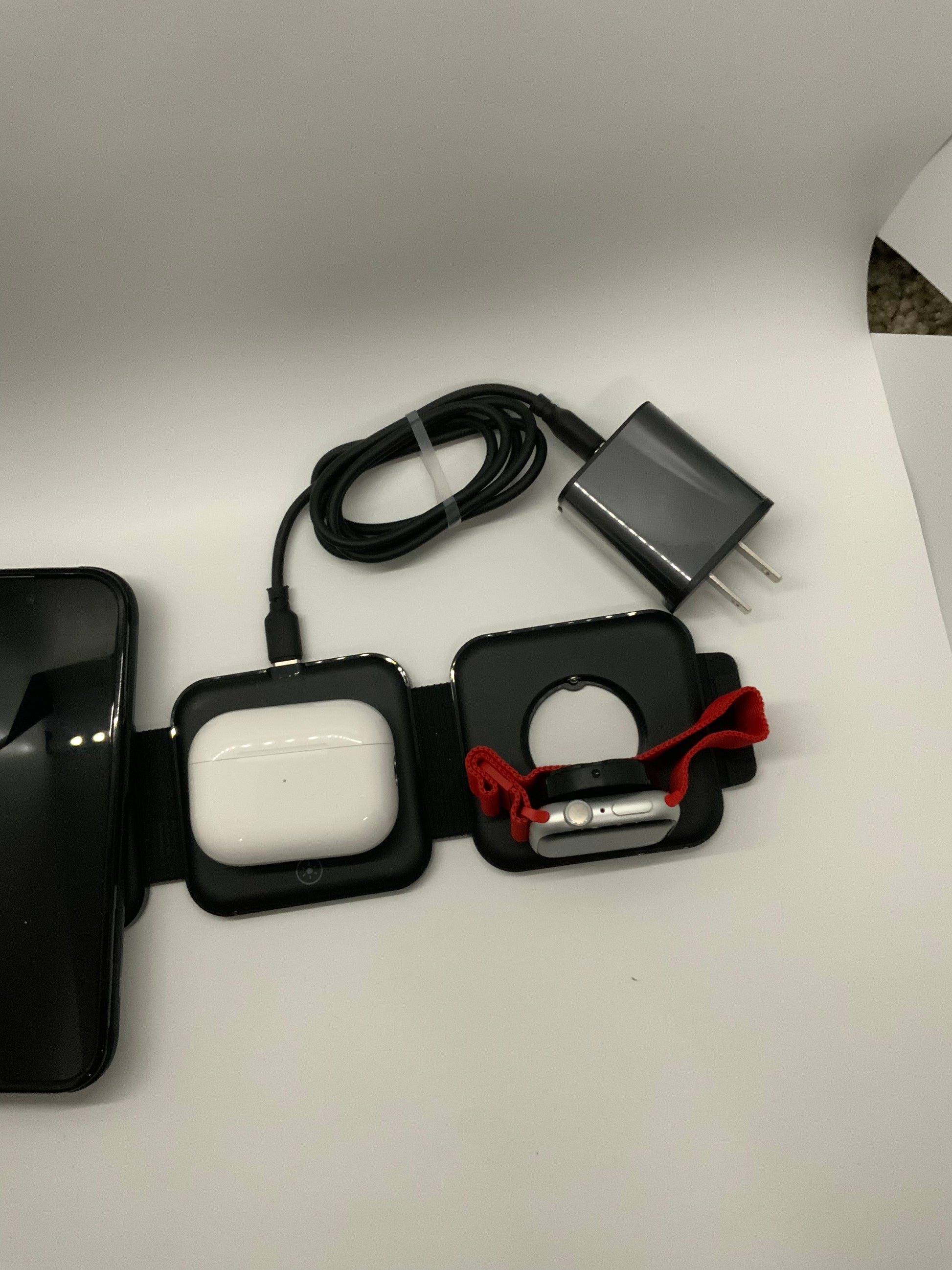 Be My AI: The picture shows several items placed on a white surface. From left to right:

1. There is a black smartphone with a reflective screen.
2. Next to the smartphone, there is a black square-shaped wireless charging pad with a white case on it that looks like it's for earbuds.
3. To the right of the charging pad, there is a smartwatch with a red woven band.
4. Above these items, there is a black cable coiled up with a silver charging block attached to it.

The background is plain white and there is a