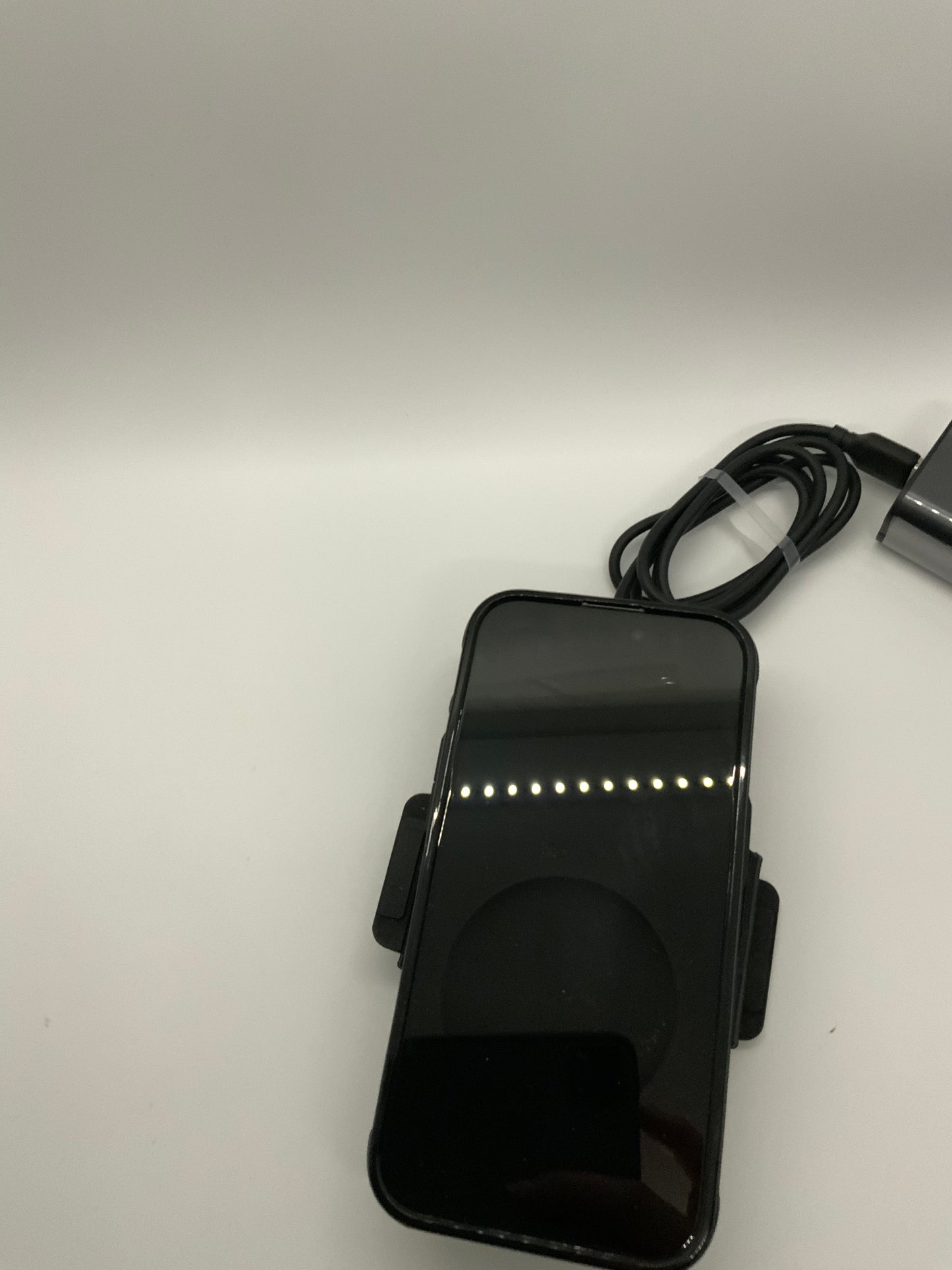 Be My AI: The picture shows a smartphone placed on a white surface. The smartphone is black and has a reflective screen. It is placed in a horizontal position and is attached to a black holder with clips on the sides. There is also a black cable coiled next to the smartphone, and it seems to be a charging cable. The background is plain white and there is a reflection of lights on the smartphone screen.