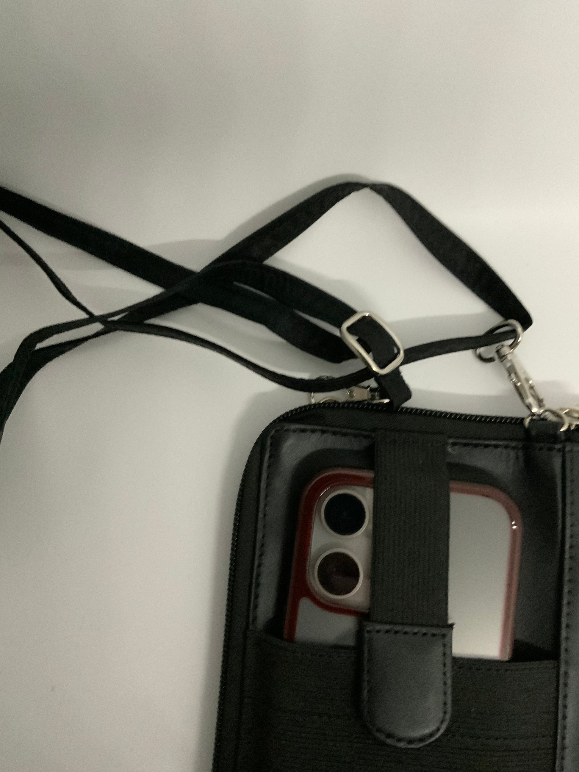 The picture shows a black wallet or small pouch with a strap. The strap is black and has a silver buckle for adjusting its length. The wallet itself has a zipper on the top and a pocket on the front with a flap that closes with a magnetic snap. Inside the front pocket, there is a smartphone with a red and white case. The smartphone's camera is visible, which has two lenses and a flash. The background is plain and white.