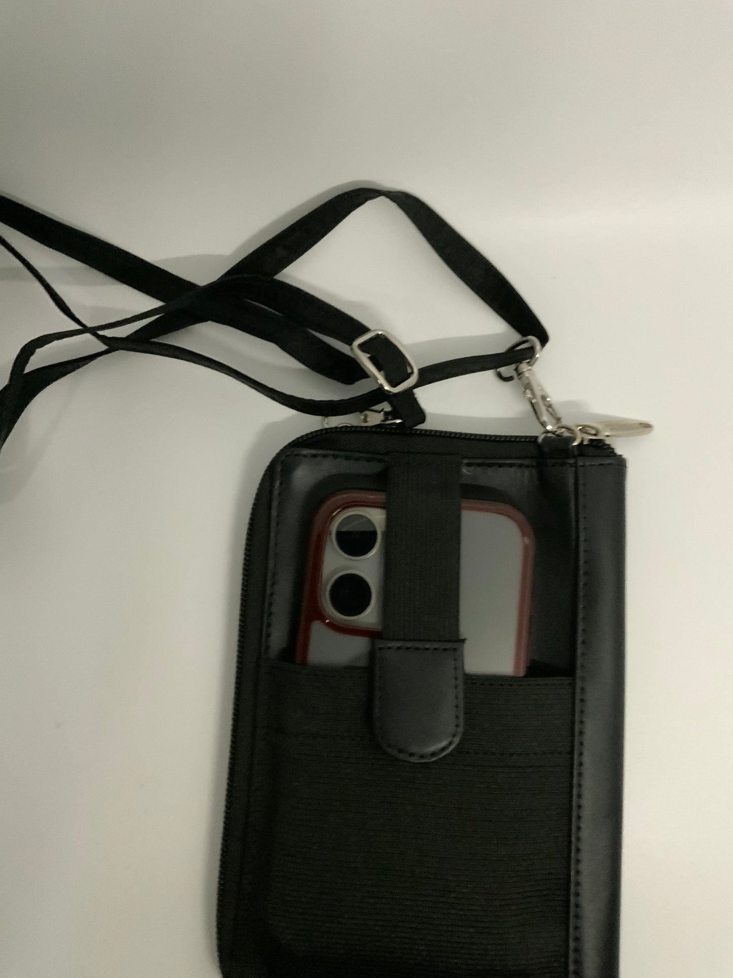 The picture shows a black phone pouch with a strap. The pouch is made of fabric and has a front pocket with a flap. Inside the front pocket, there is a smartphone with a red and white case. The phone's camera is visible. The strap of the pouch is black and has a silver buckle for adjusting its length. The strap is attached to the pouch with silver clasps. The background is plain and white.