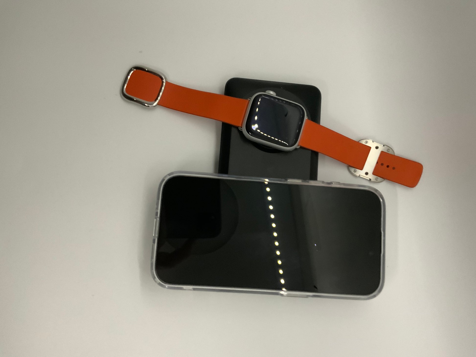 The picture shows a few electronic devices and a watch on a light-colored surface. There is a smartphone with a black screen and silver edges at the bottom. Above the smartphone, there is a smartwatch with an orange strap and a silver rectangular face. The watch is placed on top of what appears to be a black tablet or another smartphone. The background is plain and light-colored.