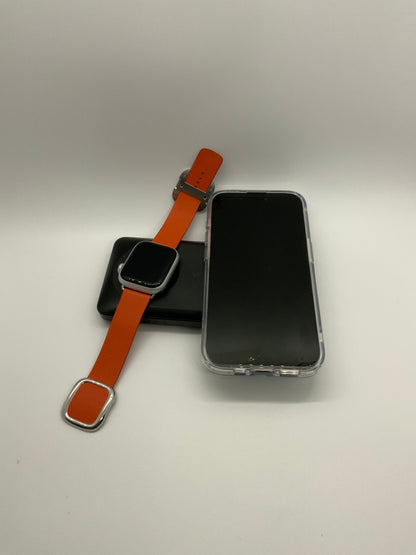 The picture shows three items placed on a white surface against a light background. 1. On the left, there is a smartwatch with a black screen and an orange strap. The strap is partially visible with a silver buckle.2. In the middle, there is a black square-shaped object, possibly a charger or a stand for the smartwatch.3. On the right, there is a smartphone with a black screen and a transparent case. The phone is lying flat with the screen facing upwards.The items are arranged in a somewha