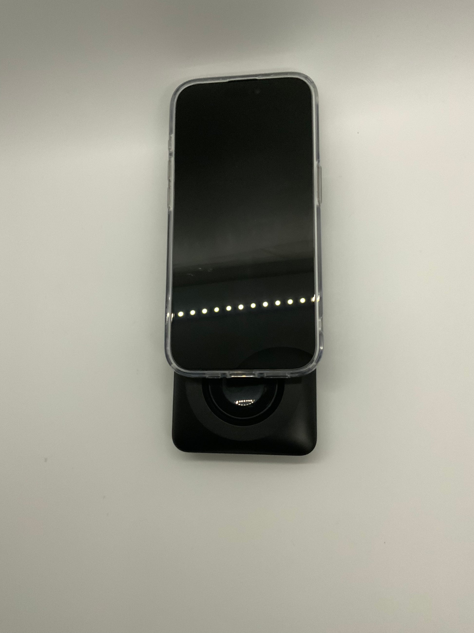 The picture shows a smartphone placed on a stand. The smartphone has a black screen and is in a vertical position. The stand is black and has a square base. The background is plain and light-colored. The phone is not turned on, so nothing is displayed on the screen.