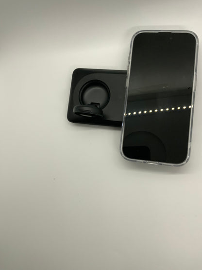 The picture shows two objects on a white surface. On the right side, there is a smartphone with a black screen and a silver edge. The phone is lying with its screen facing upwards. On the left side, there is a black rectangular object with a circular cutout in the middle. It looks like a phone holder with a ring that can be used to grip the phone. The background is plain white.