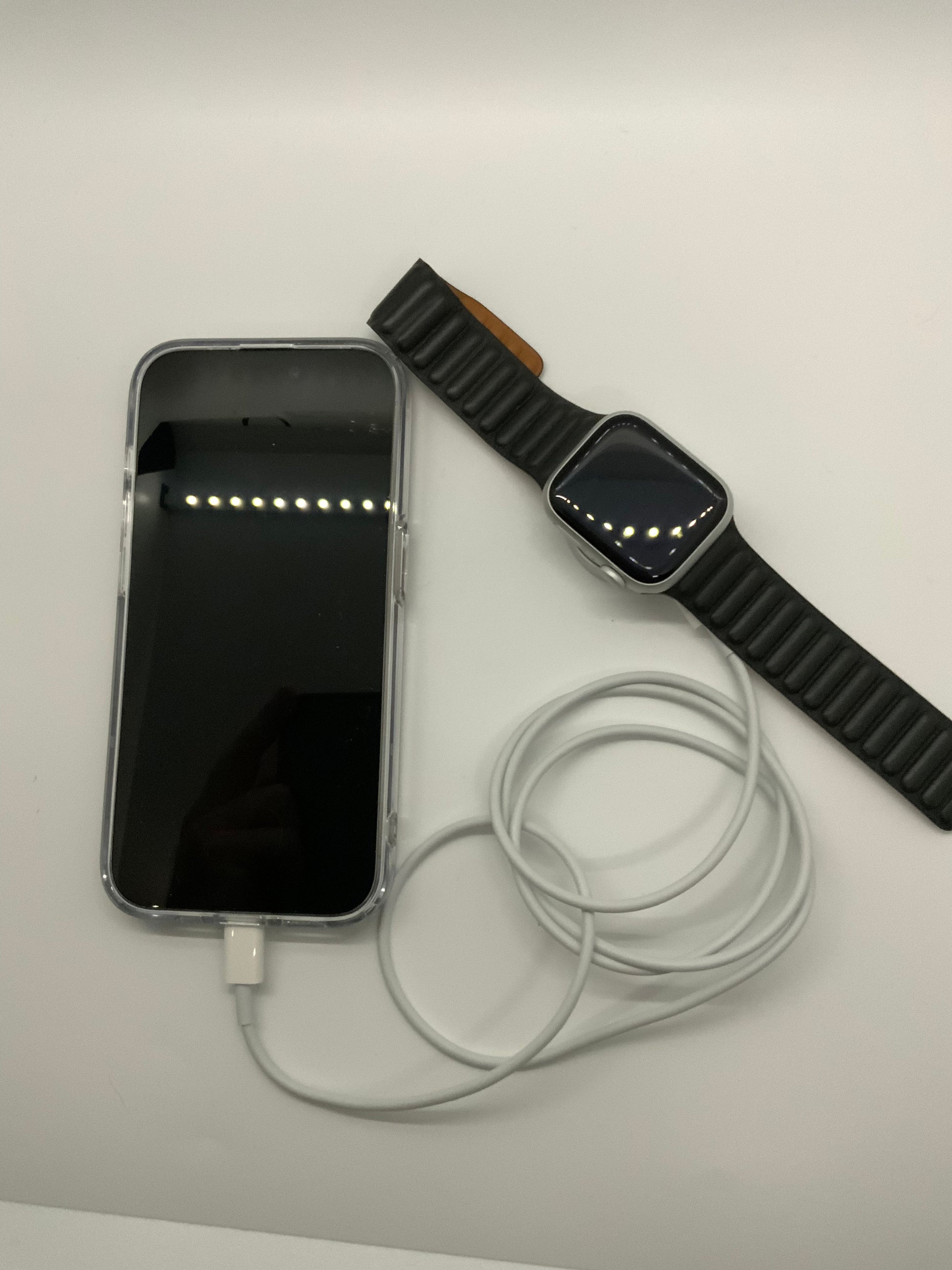 Be My AI: The picture shows a smartphone, a smartwatch, and a charging cable on a white surface. The smartphone is on the left side of the image with its screen facing up. The screen is black and reflects some lights. The charging cable is plugged into the smartphone and is coiled up next to it. The smartwatch is on the right side of the image with its screen facing up as well. The smartwatch has a black strap and its screen is also black, reflecting some lights.