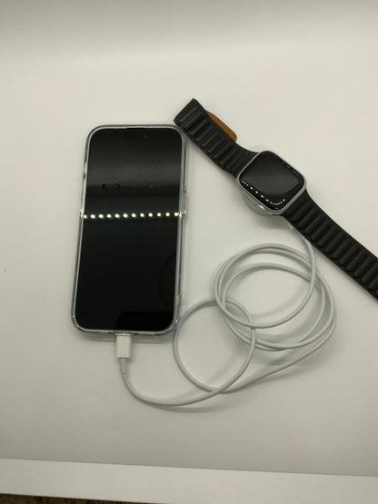 The picture shows a smartphone, a smartwatch, and a charging cable on a white surface. The smartphone is on the left side of the image and has a black screen with a white charging cable plugged into it. The cable is coiled loosely next to the phone. On the right side of the image, there is a smartwatch with a black band. The watch face is black and the band has a ridged texture. The background is plain white.