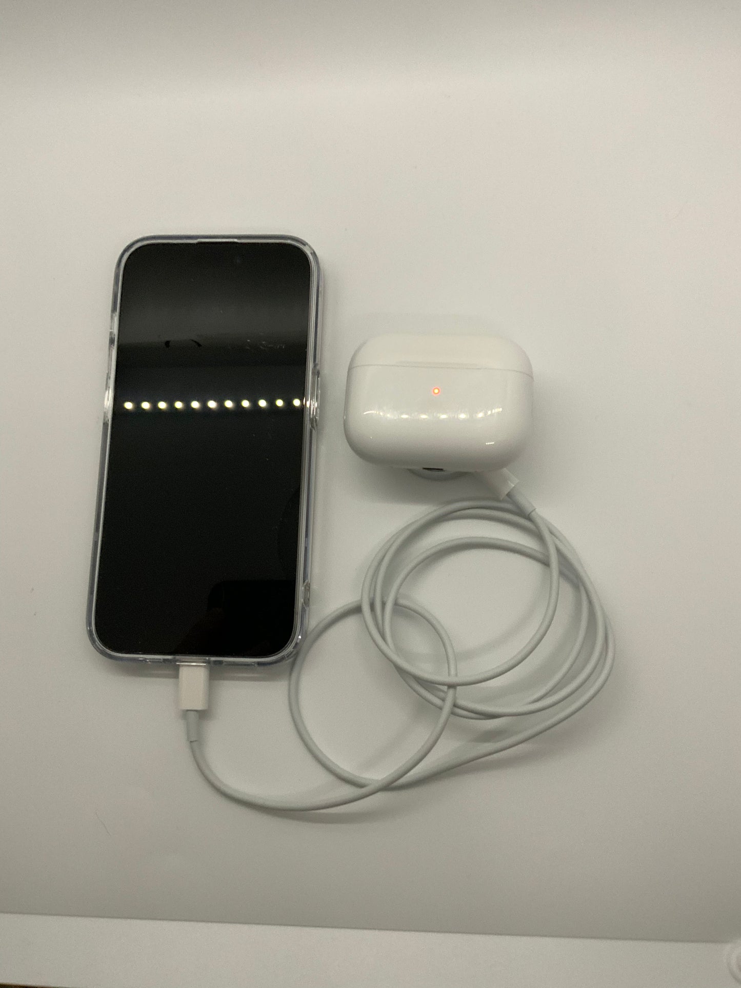 The picture shows a smartphone with a white charging cable plugged into it. Next to the smartphone, there is a white case for wireless earbuds with a small red light on it. The case also has a charging cable attached to it. Both the smartphone and the earbud case are placed on a white surface.