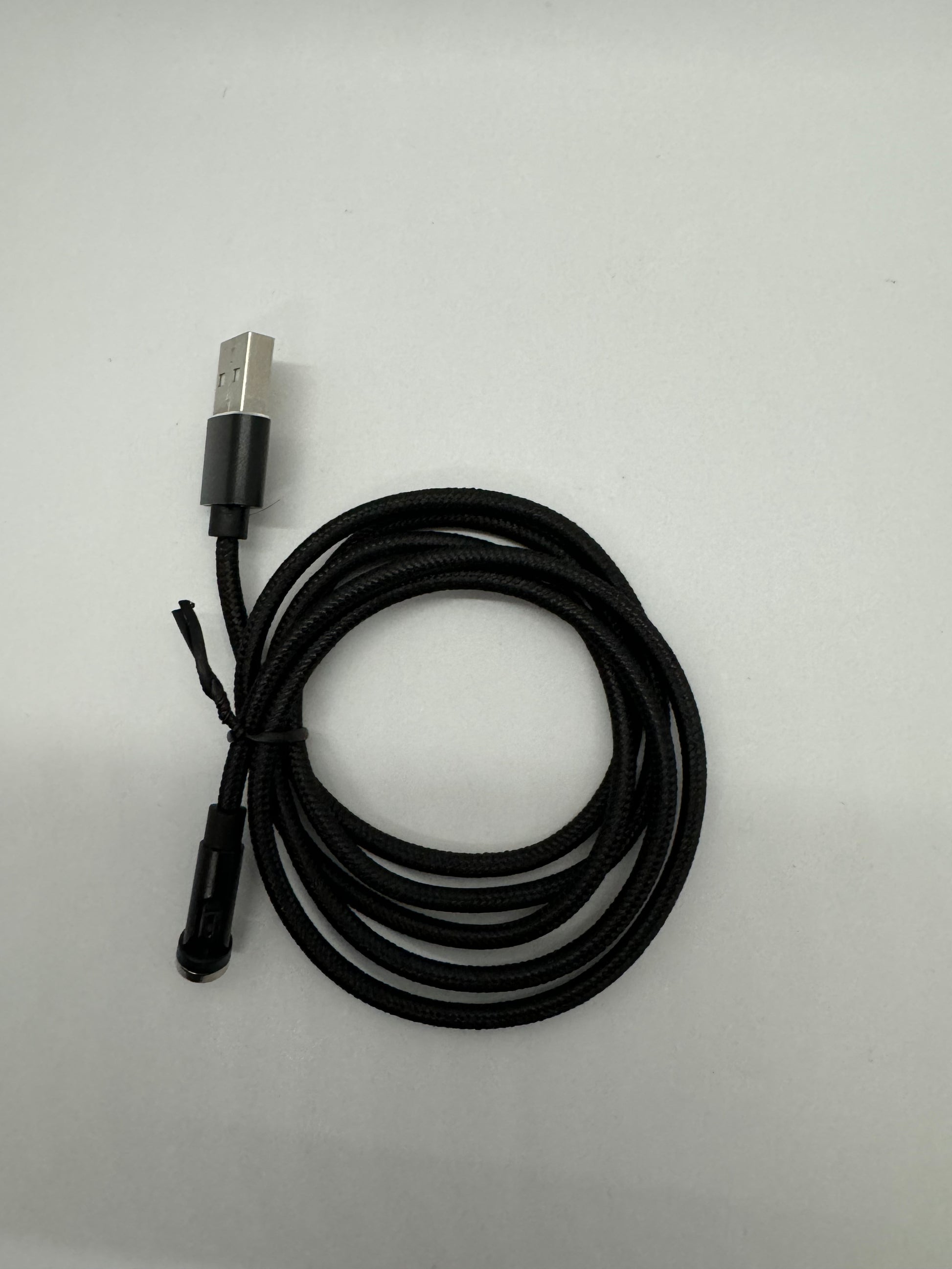 The picture shows a black USB cable on a white background. The cable is coiled up and has a braided texture. One end of the cable has a standard USB connector, which is silver with a black casing. The other end of the cable is not fully visible, but it appears to be a smaller connector, possibly a micro USB, and it is also black.