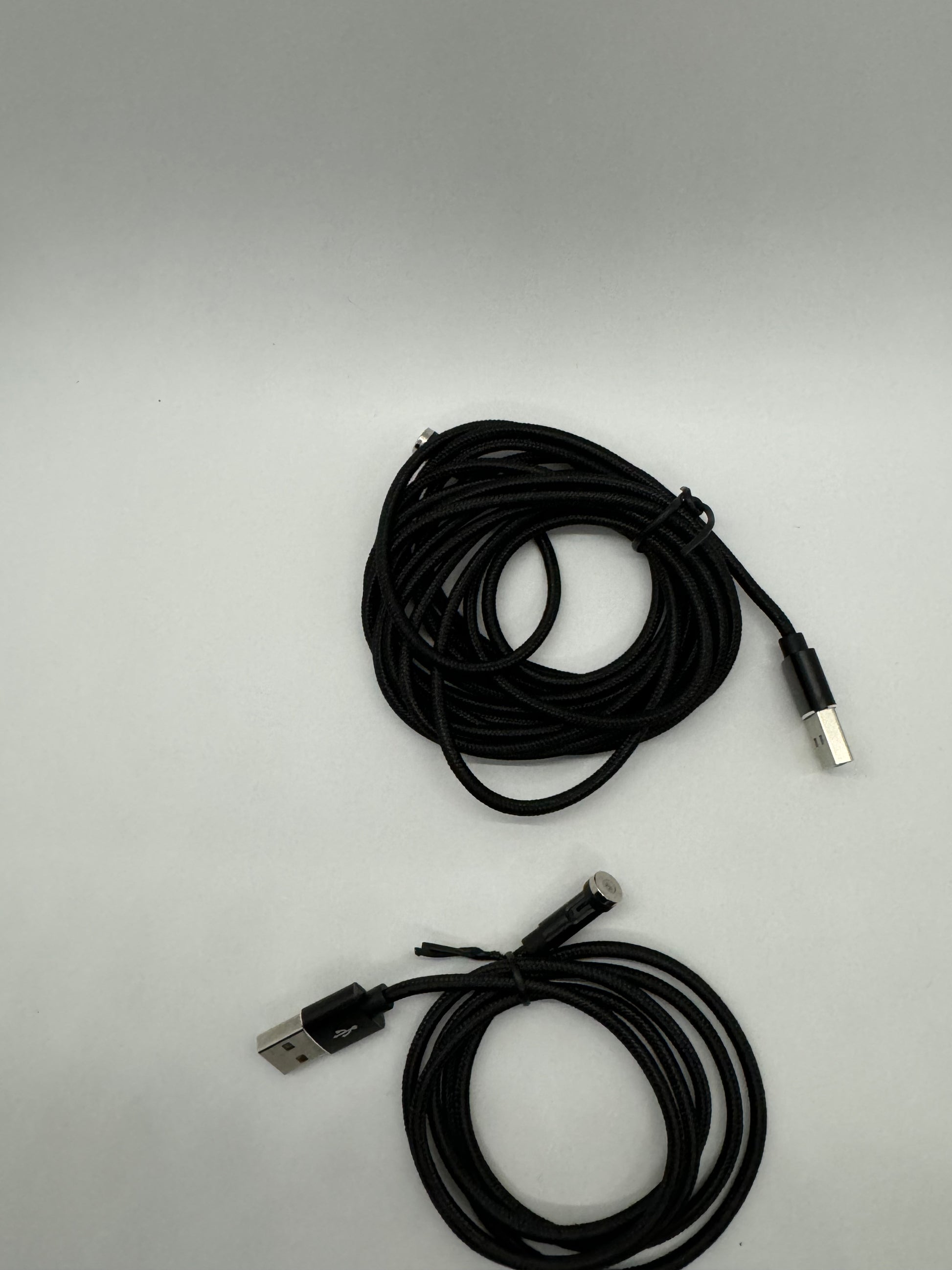 The picture shows two black cables on a white background. Both cables are coiled. One of the cables has a USB connector on one end and a lightning connector on the other end. The other cable has a USB connector on one end and what appears to be a micro USB connector on the other end. The cables appear to be made of a braided material.