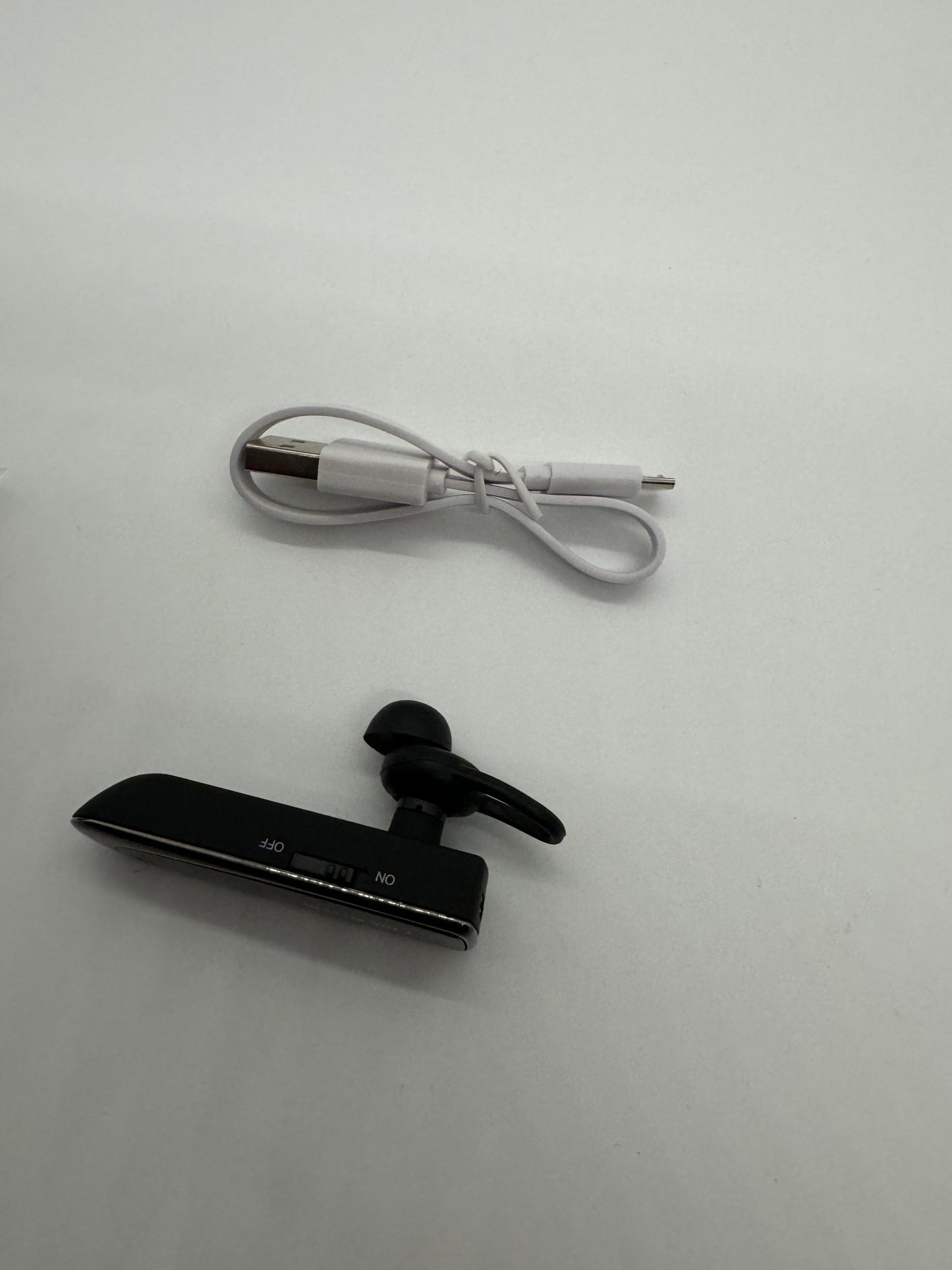 The picture shows two items on a white background. The first item is a white cable, possibly a charging cable. It is coiled up and has a USB connector on one end and a smaller connector, possibly micro USB, on the other end.The second item is a black Bluetooth earpiece. It has a clip on one side, an earbud, and a small rectangular body. On the body, there are words that appear to be "ON" and "OFF" indicating the switch positions, but it is mirrored.