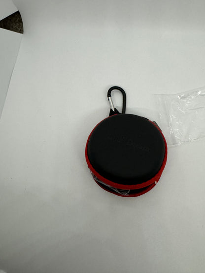 The picture shows a small round case with a black carabiner attached to it. The case appears to be black with a red zipper. It is placed on a white surface. There is also a clear plastic bag partially visible in the picture.