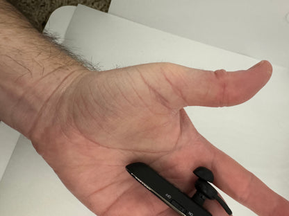 The picture shows a person's hand, specifically the palm side of their thumb and part of the wrist. The hand is resting on a white surface. The skin on the hand is light and there are some visible hairs on the wrist. The thumb is slightly curved. Near the base of the thumb, there is a black object which appears to be a small electronic device, possibly a USB or a small remote. It has two buttons on it, one labeled "NO" and the other labeled "CH". There is also a small black attachment on the top o