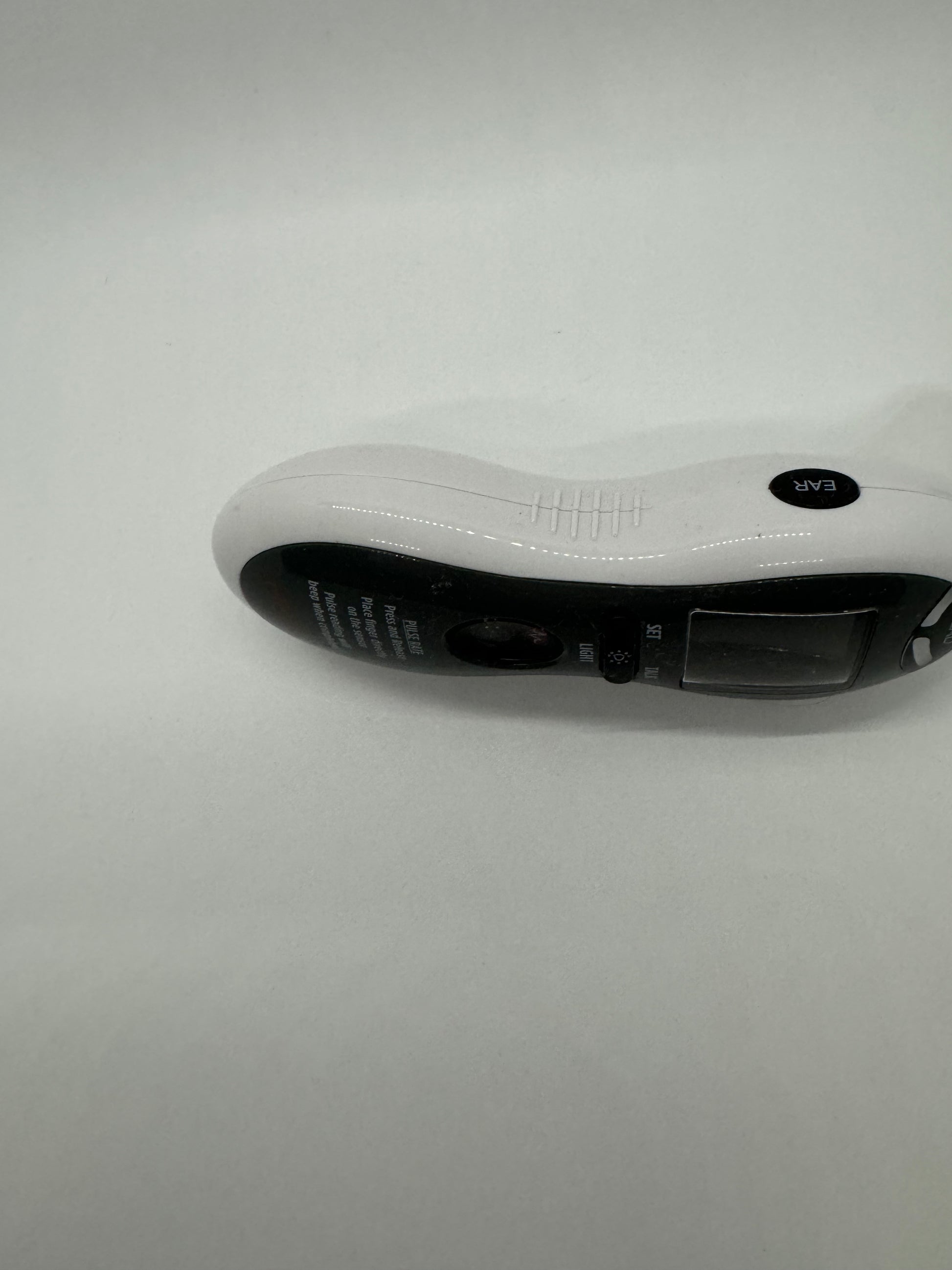 The picture shows a white object on a plain white background. The object appears to be a small electronic device, possibly a remote control or a handheld scanner. It has an ergonomic design with a curved shape to fit comfortably in the hand. The top part of the device is white with a textured grip and a small black circular button with the letters "EVA" on it. The bottom part of the device is black with some text, but it's not fully visible in the picture.