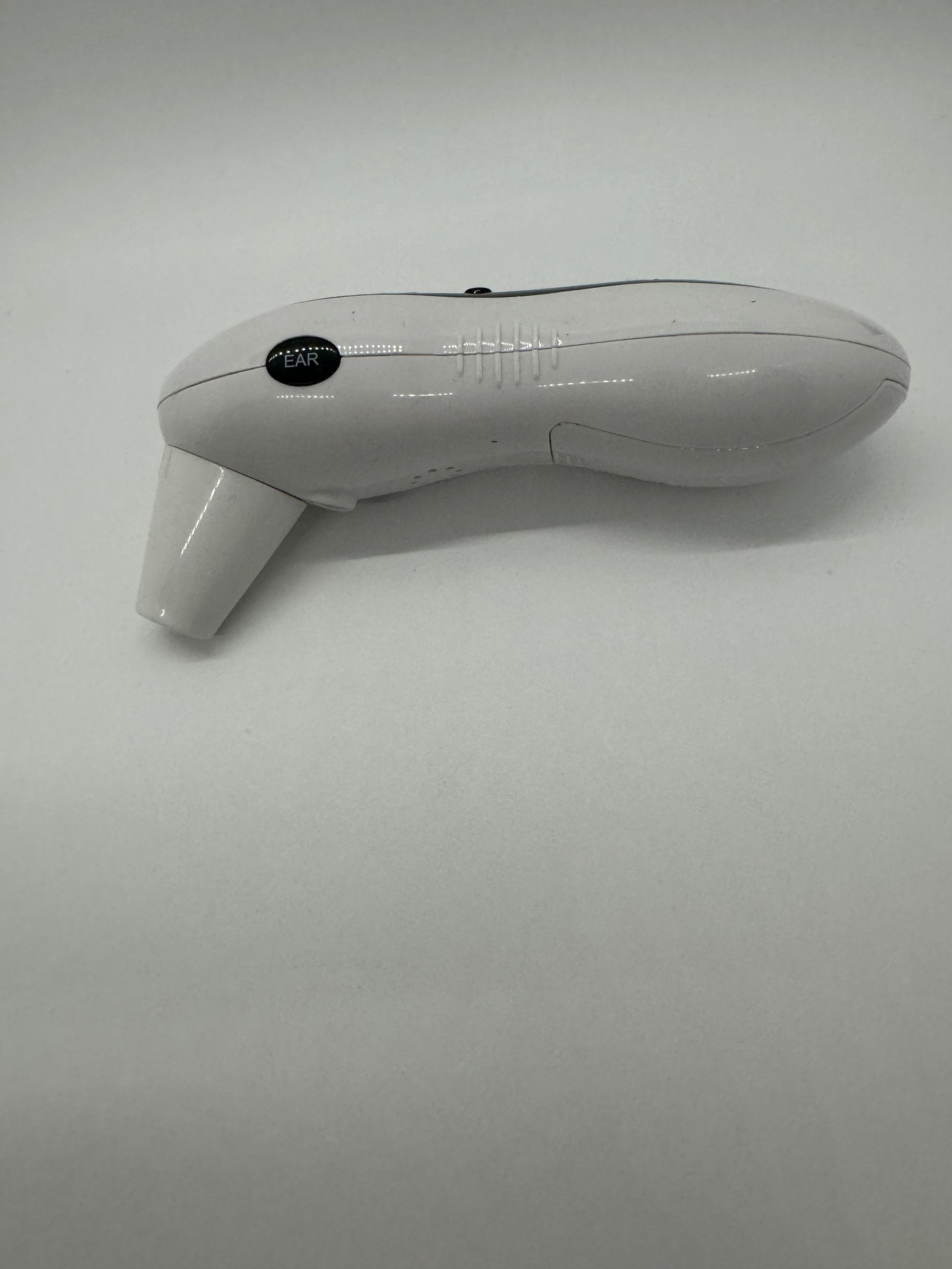 The picture shows a device that appears to be an ear thermometer. It is placed on a plain white background. The device is light grey in color and has an ergonomic design, shaped to fit comfortably in the hand. The top part of the device is elongated and curves down into a pointed tip, which is likely the part that is inserted into the ear. On the side of the device, there is a small round black button with the word "EAR" written in white letters. There is also a grip texture on the handle for easy