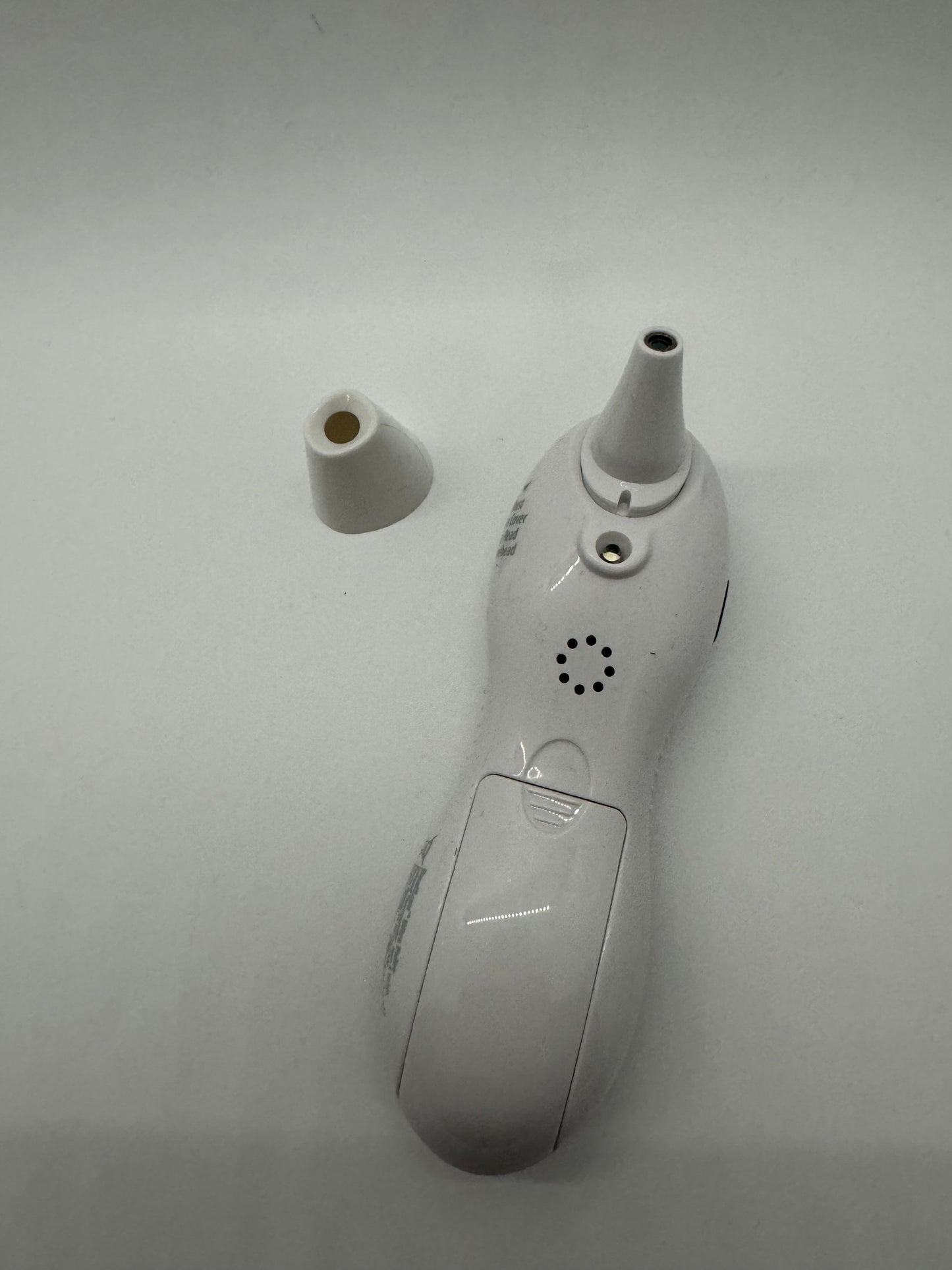 The picture shows two objects on a white background. One object is a small white cap or lid, which is cylindrical in shape with a hollow center. The other object appears to be a white electronic device, possibly a thermometer. The device has a narrow, elongated top with a small opening, similar to the mouth of a bottle. The body of the device is somewhat oval in shape with a clear plastic section in the middle that might be a display screen. There are also small holes near the top of the device, w